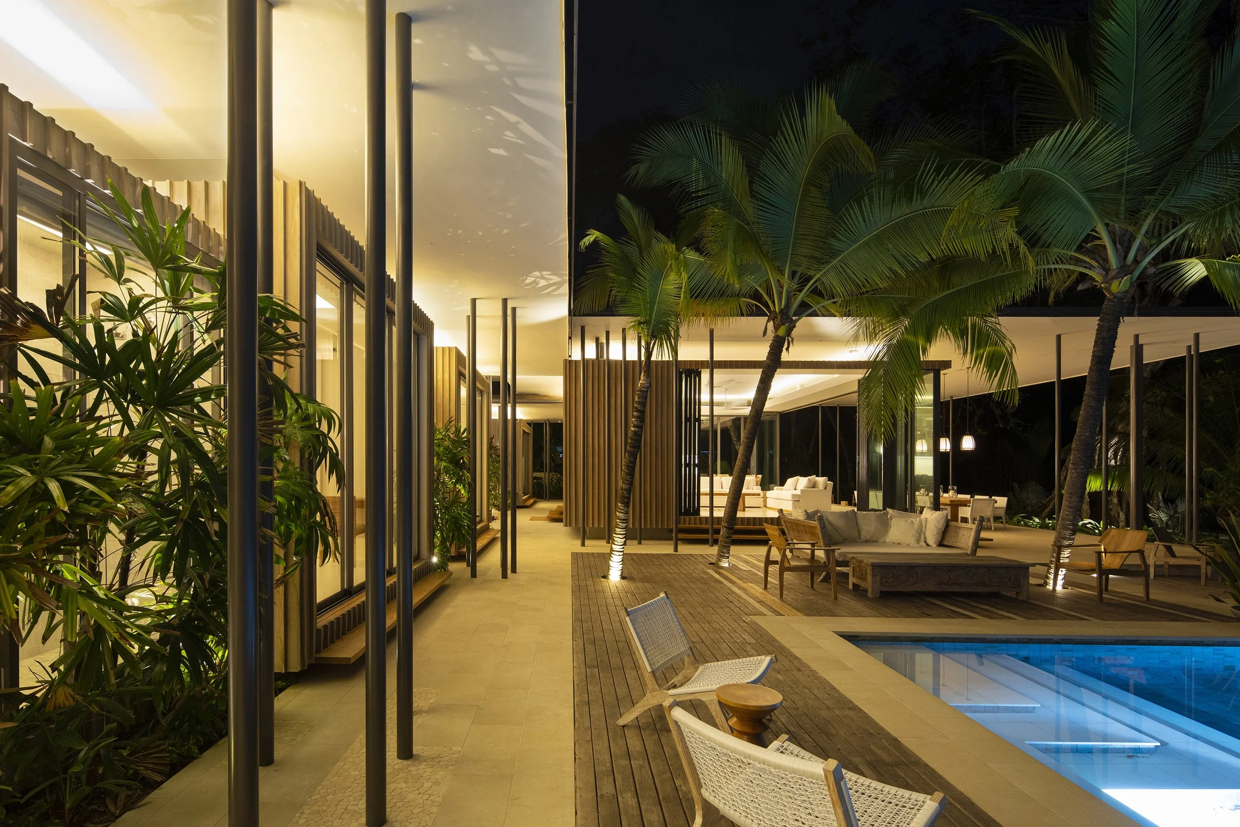 Modern outdoor patio area at night with tropical plants, chairs, and a swimming pool, illuminated by warm lighting.