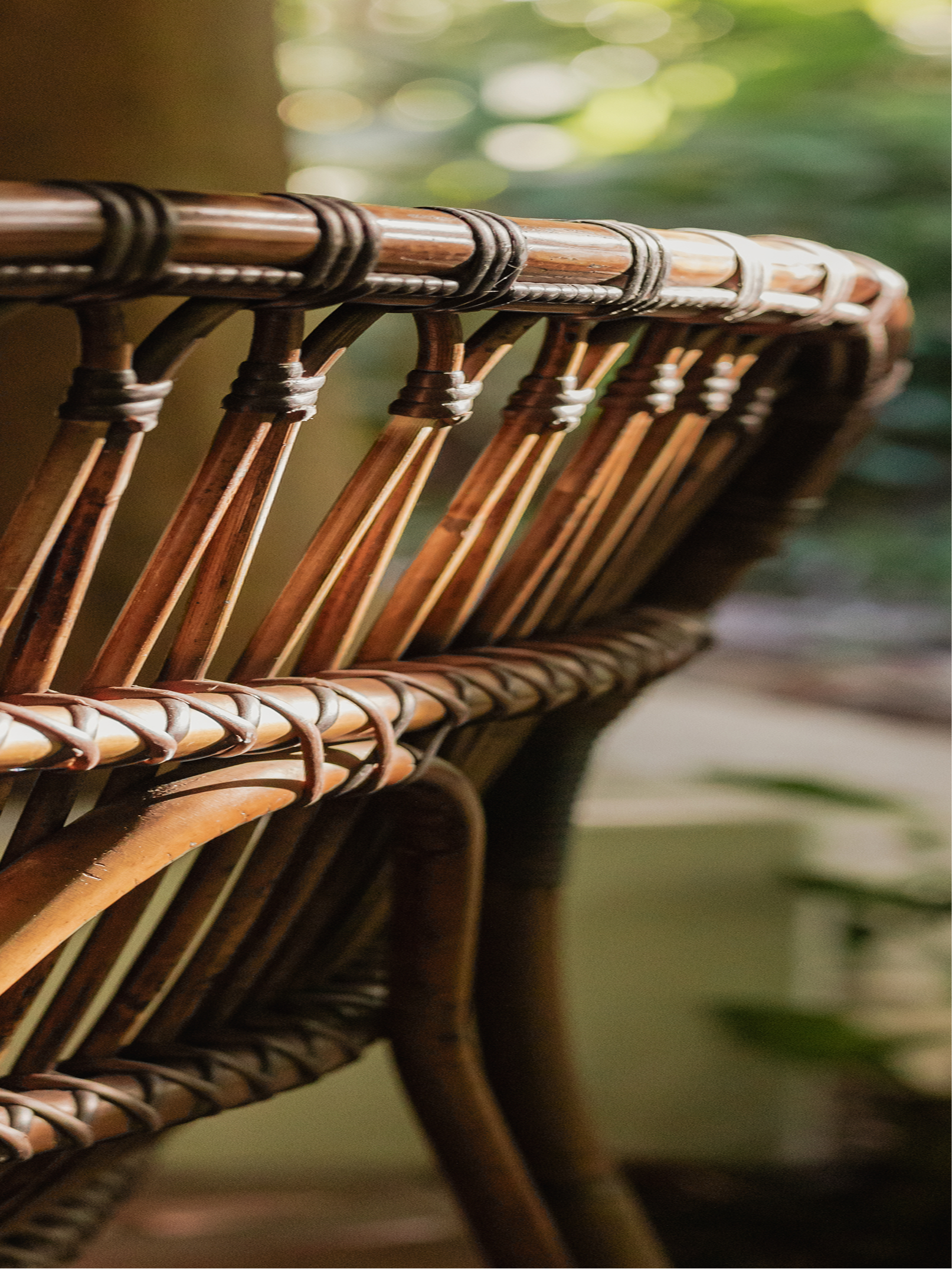 Close-up of a rattan chair with a wooden top rail and vertical supports, with a blurred green and white background.