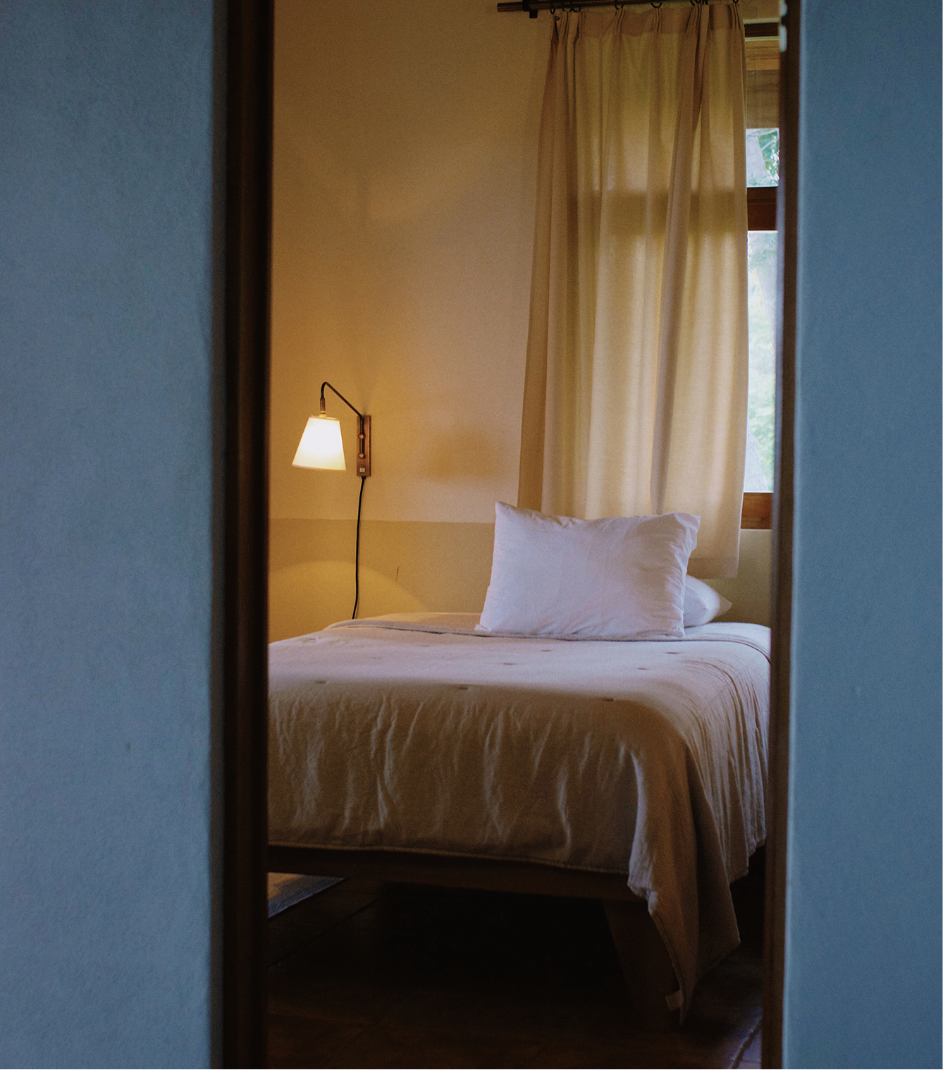 A cozy bedroom seen through a mirror reflection, featuring a bed with white sheets and pillow, a wall-mounted reading lamp, and cream-colored curtains over a window with a view of trees outside.