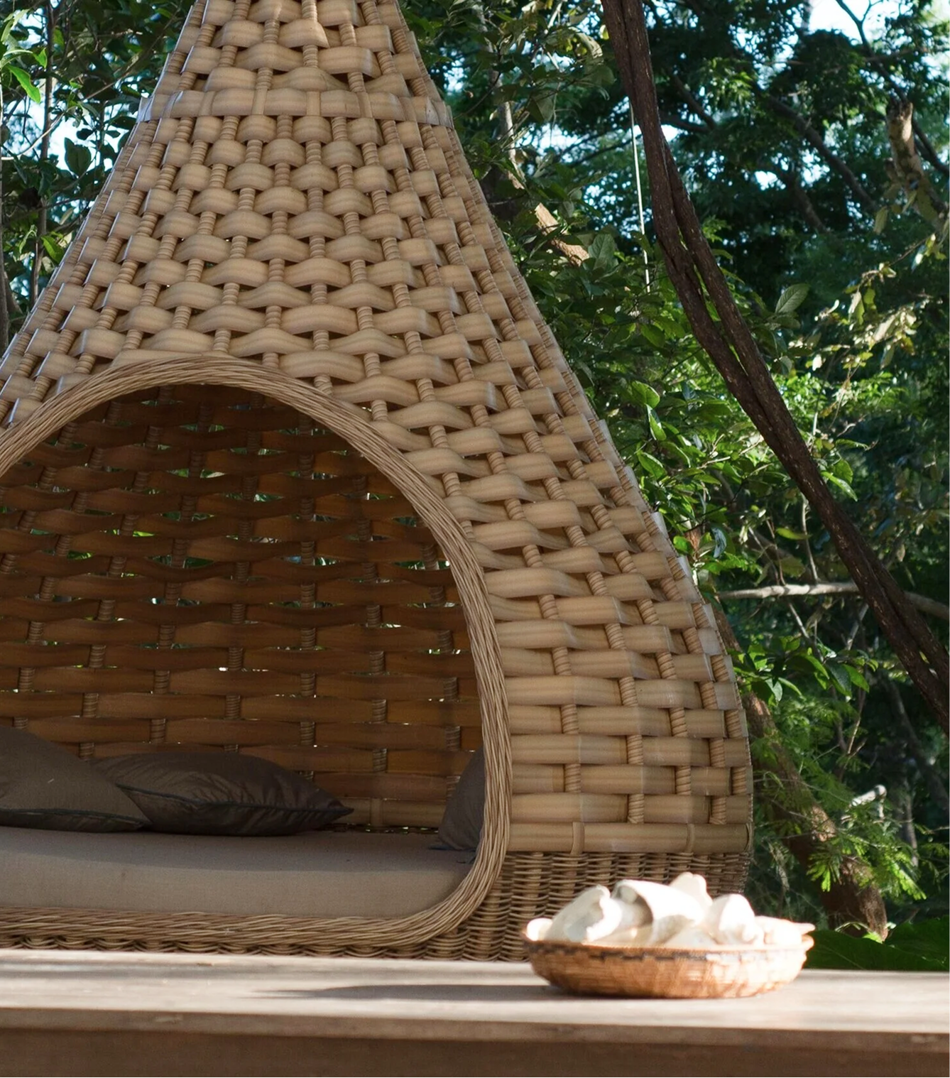 A woven outdoor cabana with a cushioned seat and pillows, set in a lush green natural environment, with a small bowl of seashells on a wooden surface in the foreground.