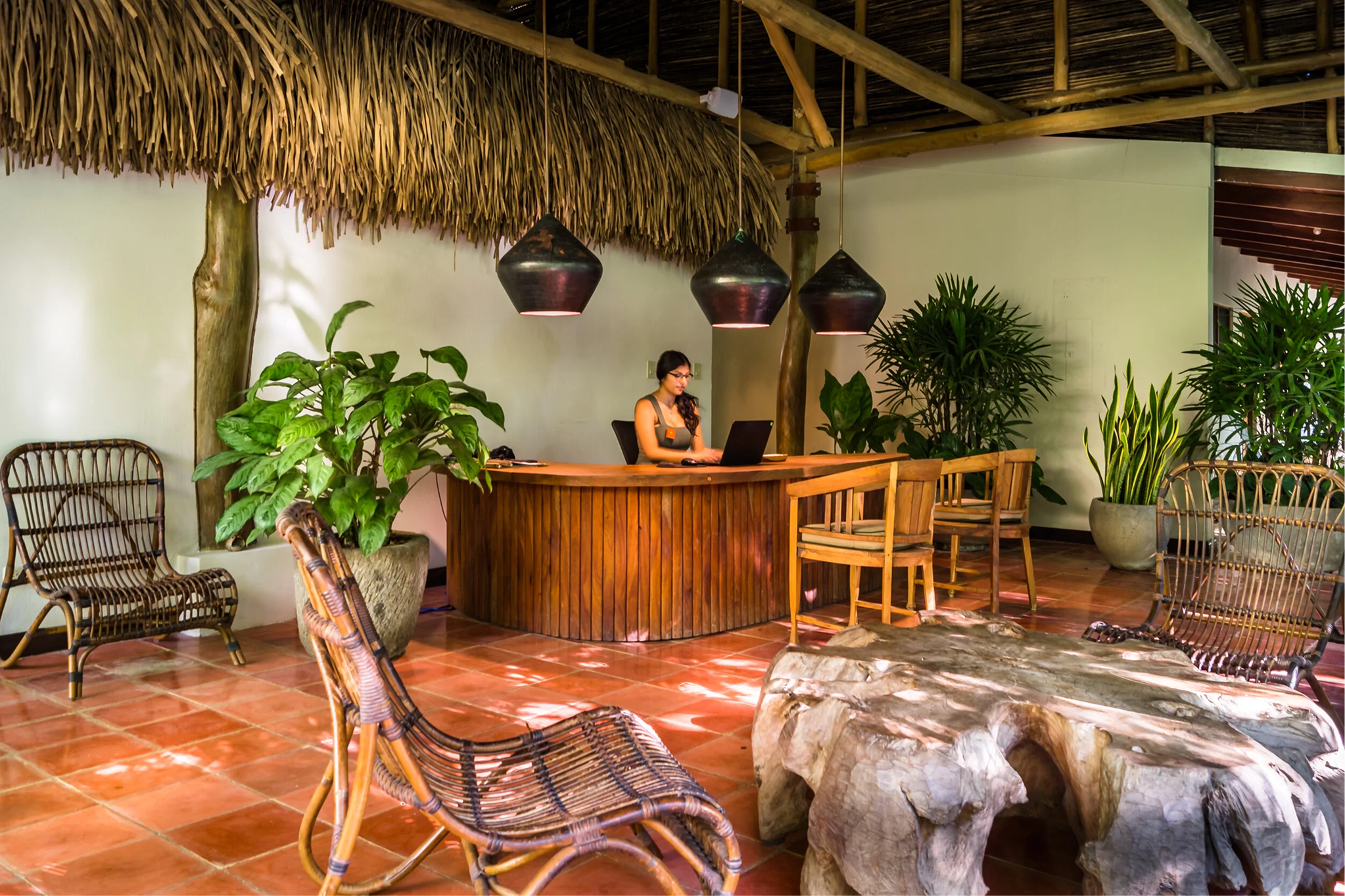 A tropical-themed interior with a thatched roof, bamboo beams, and lush green potted plants. A woman works on a laptop behind a wooden reception desk under three black pendant lights. Wicker chairs surround a rustic stone table in the foreground.