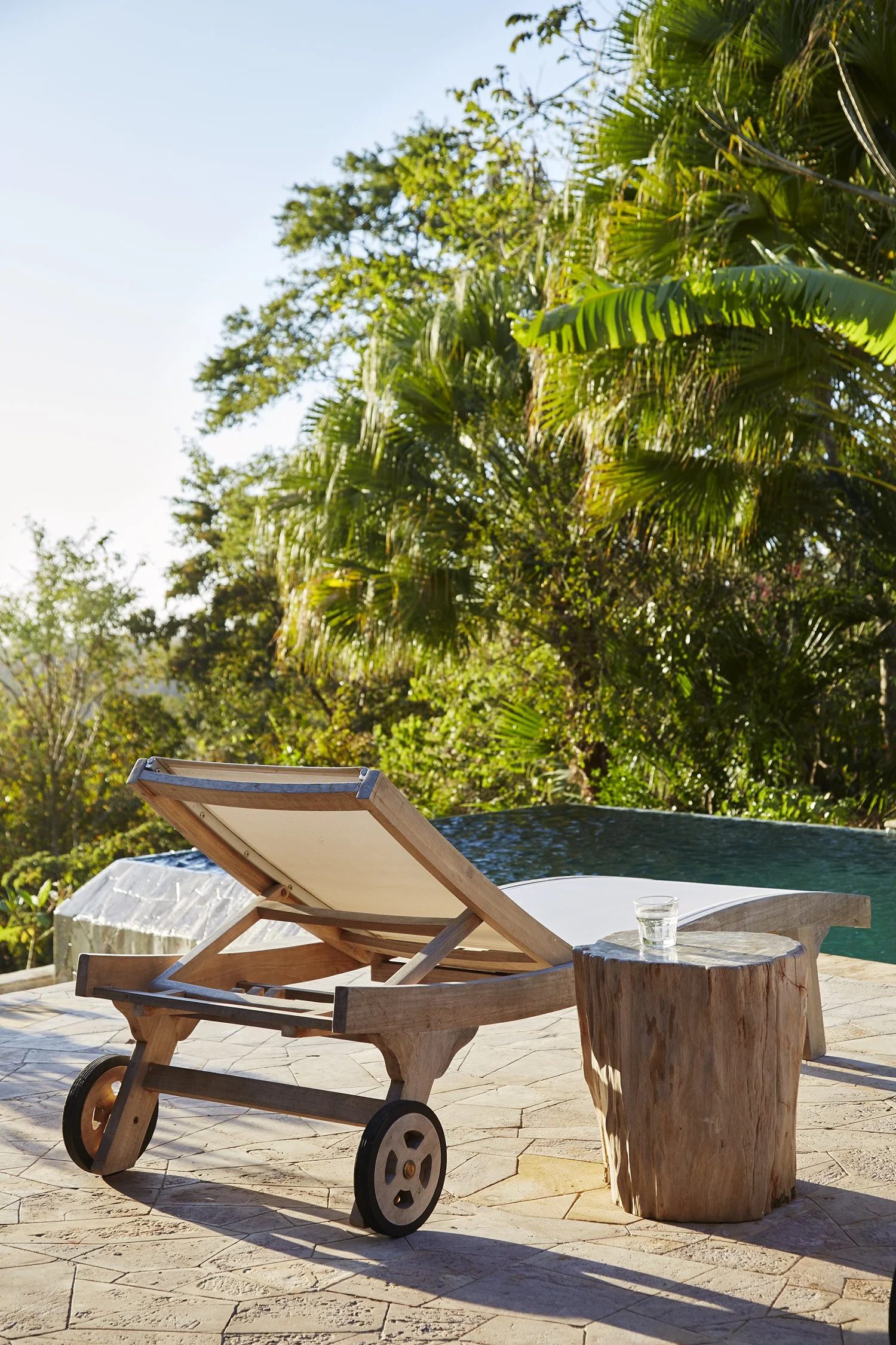 A wooden outdoor lounge chair with wheels, a cylindrical wooden side table with a glass of water, and a wooden table, set on a stone patio. In the background, lush green trees and bushes are visible, with some sunlight and a clear blue sky.