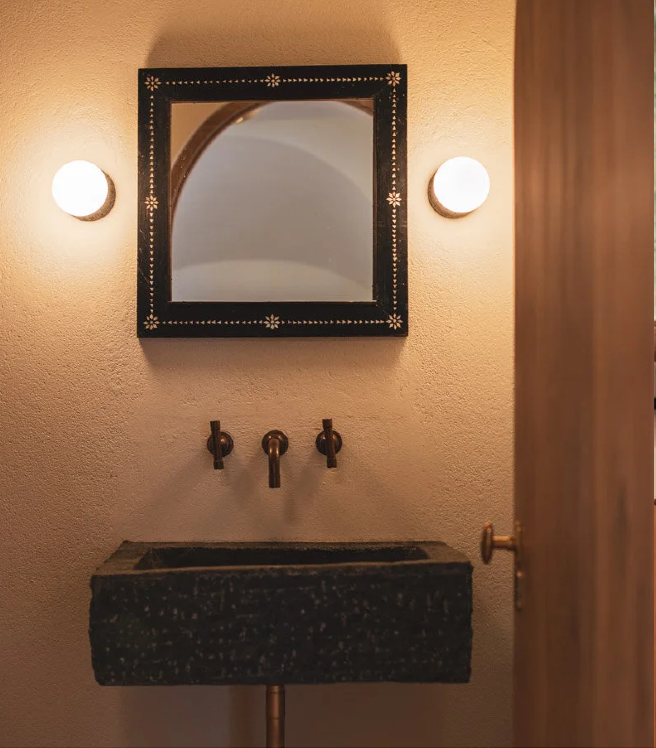 Wall-mounted mirror with black decorative frame, flanked by two round wall lights, above a granite countertop with brass faucet handles.