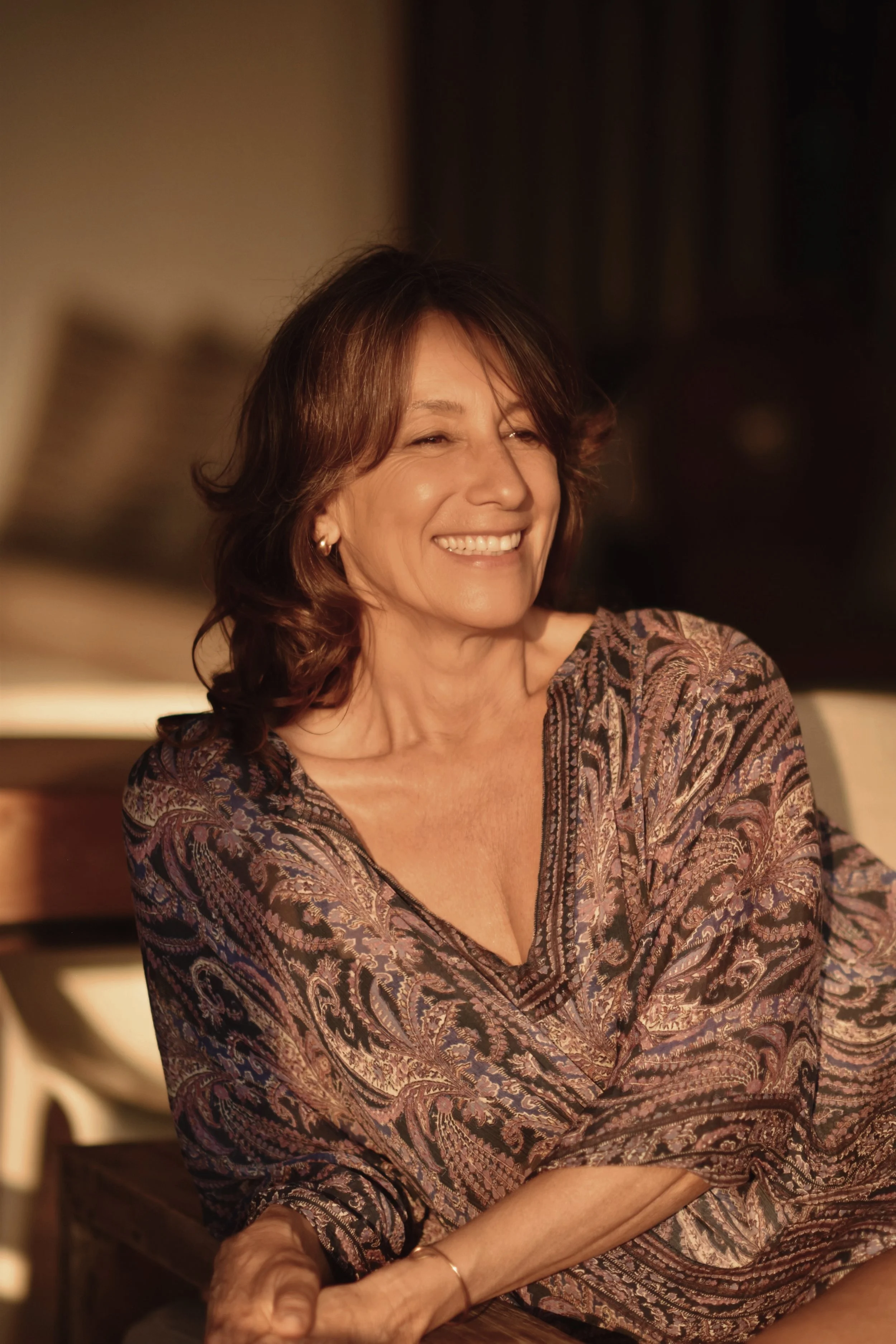 A woman with brown hair and a broad smile sitting at a table, wearing a patterned blouse in warm lighting.