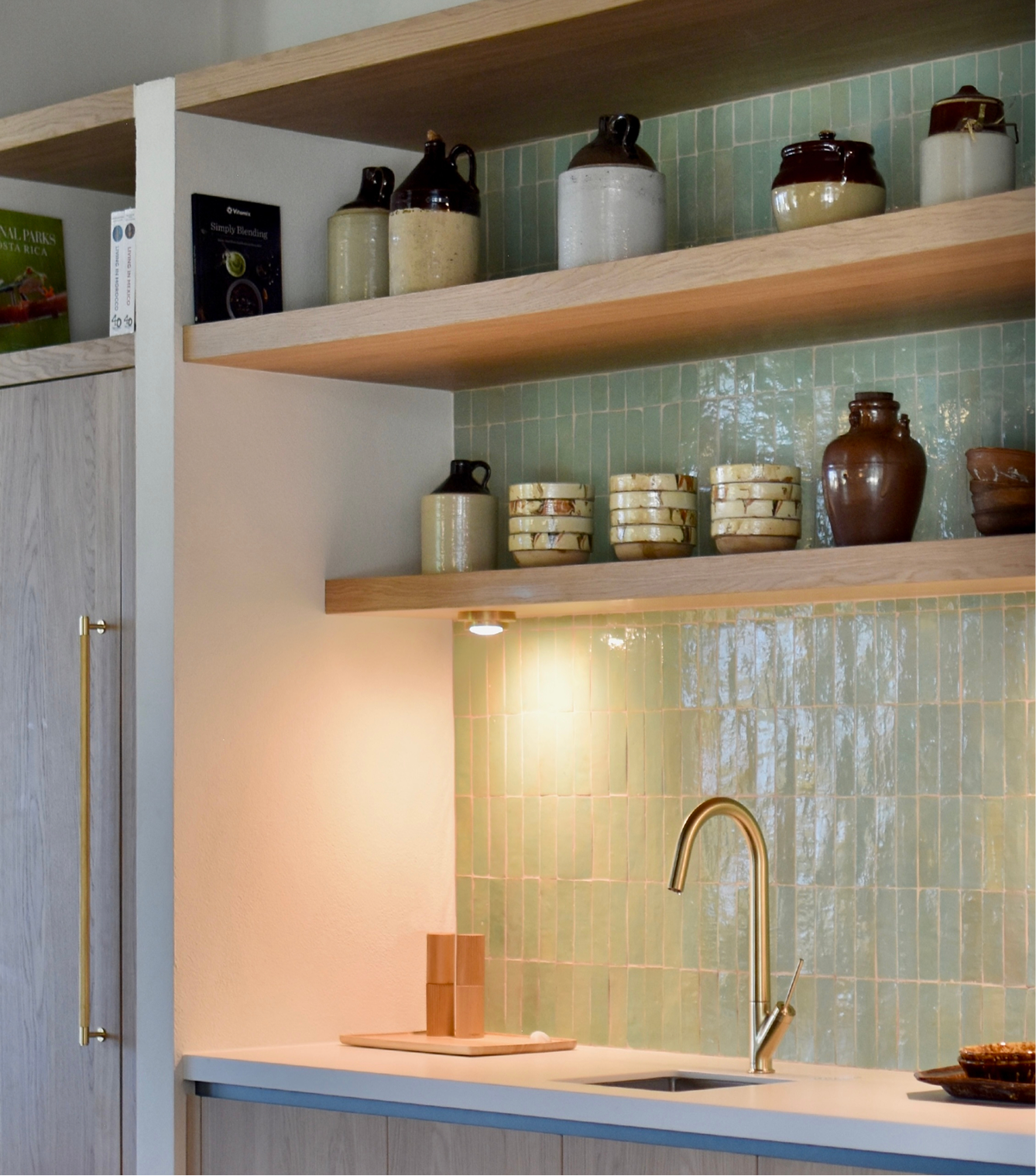 Open kitchen with light wood and white cabinetry, green tiled backsplash, brass faucet, and wooden shelves holding ceramic jars and bowls.