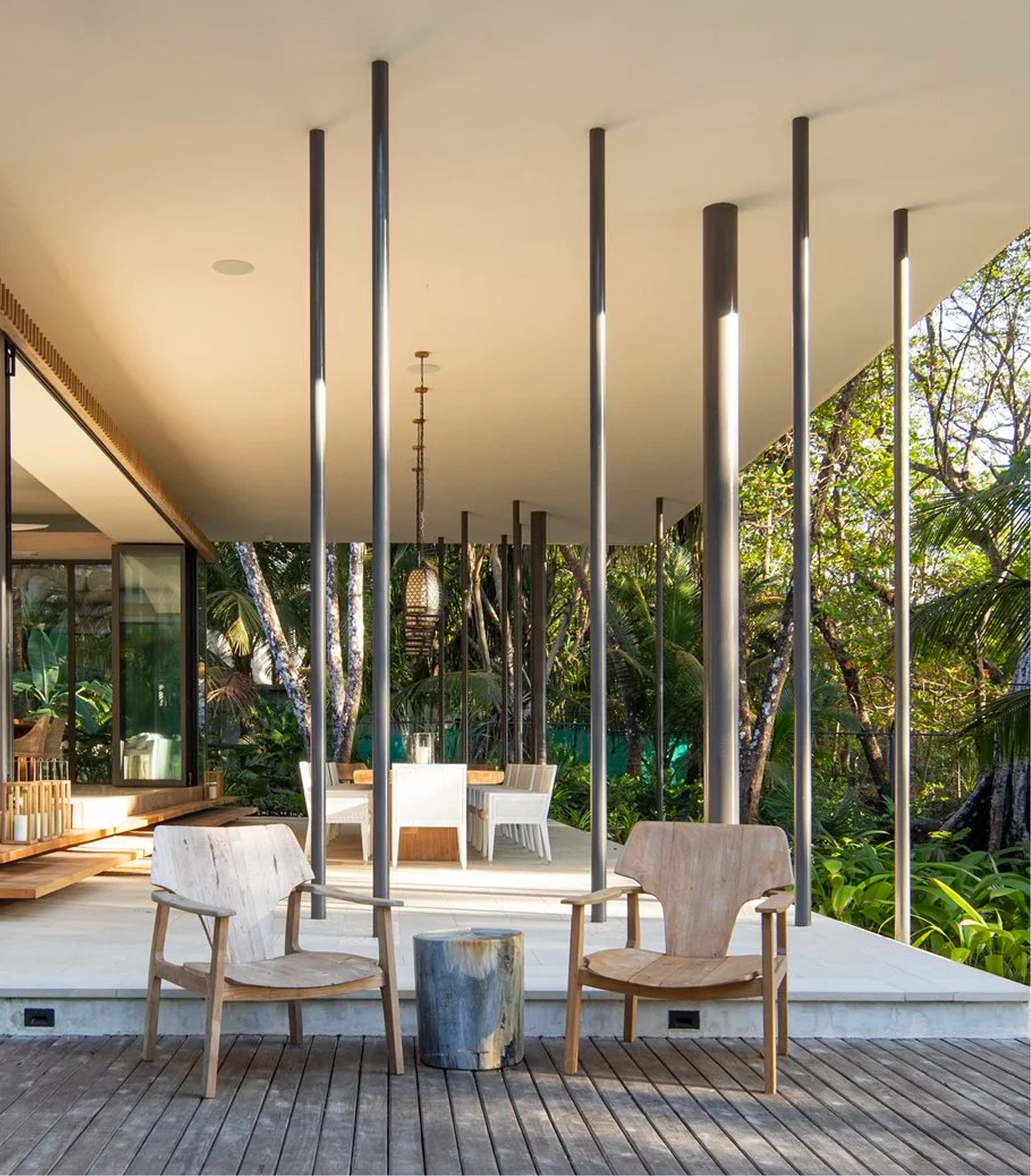 A covered outdoor patio with a white ceiling, supported by vertical black poles. There are wooden chairs and a small side table on the wooden deck, and lush green trees and plants in the background.