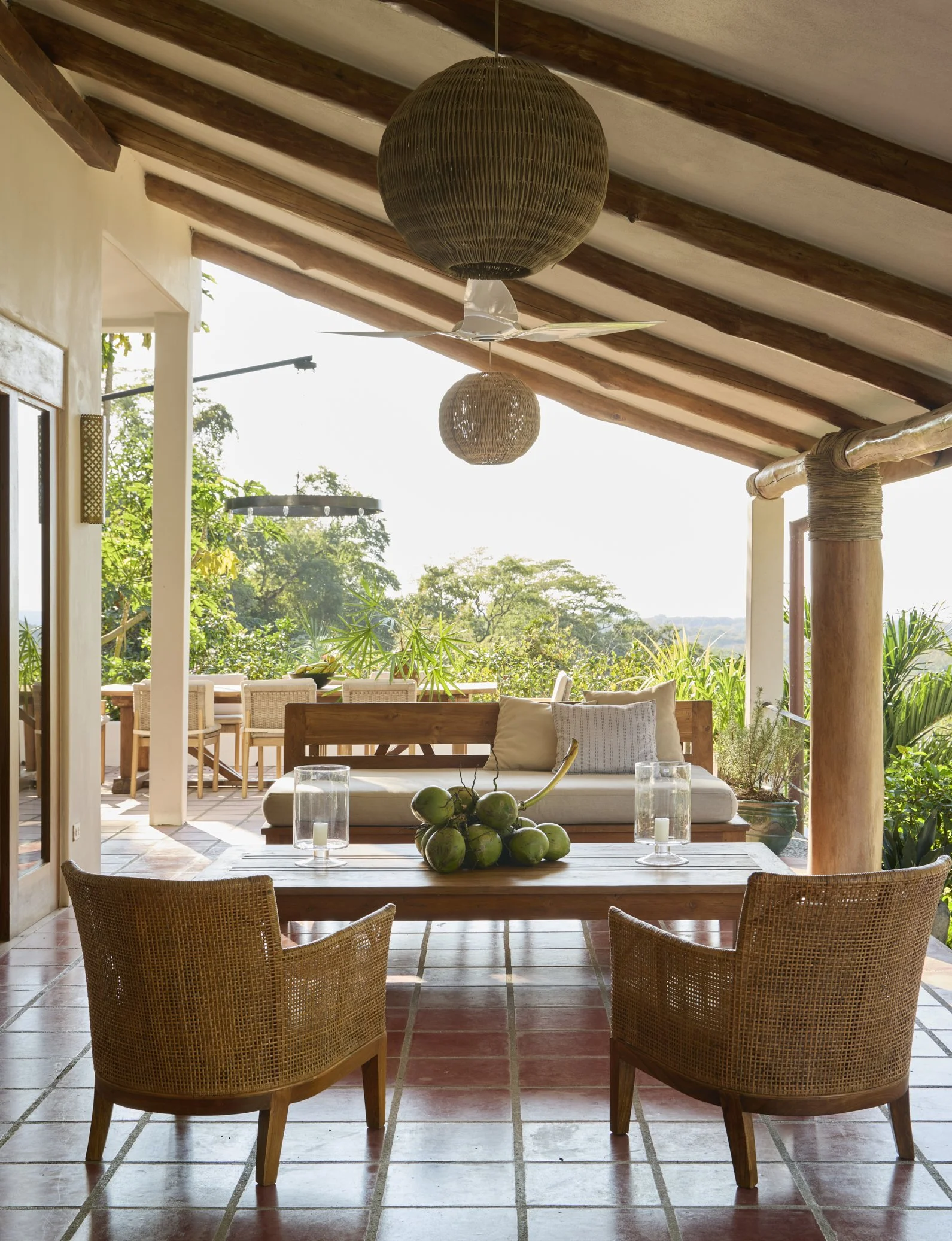 Indoor patio area with wicker chairs around a wooden table, large green coconuts in the center, and glass candle holders. Open view to lush outdoor greenery and trees, wooden ceiling beams, hanging woven light fixtures, and a white ceiling fan.