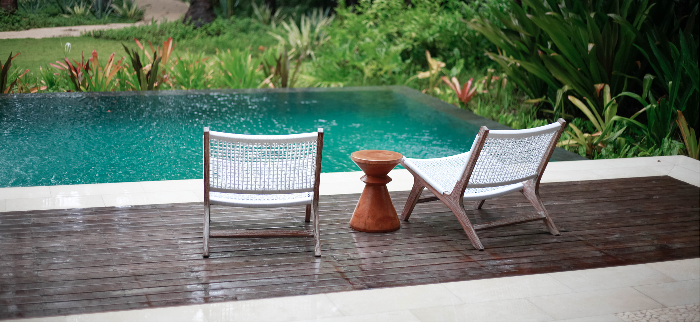 Two white modern chairs with wooden frames and a small wooden table between them sit on a wet wooden deck overlooking a swimming pool surrounded by lush green plants.