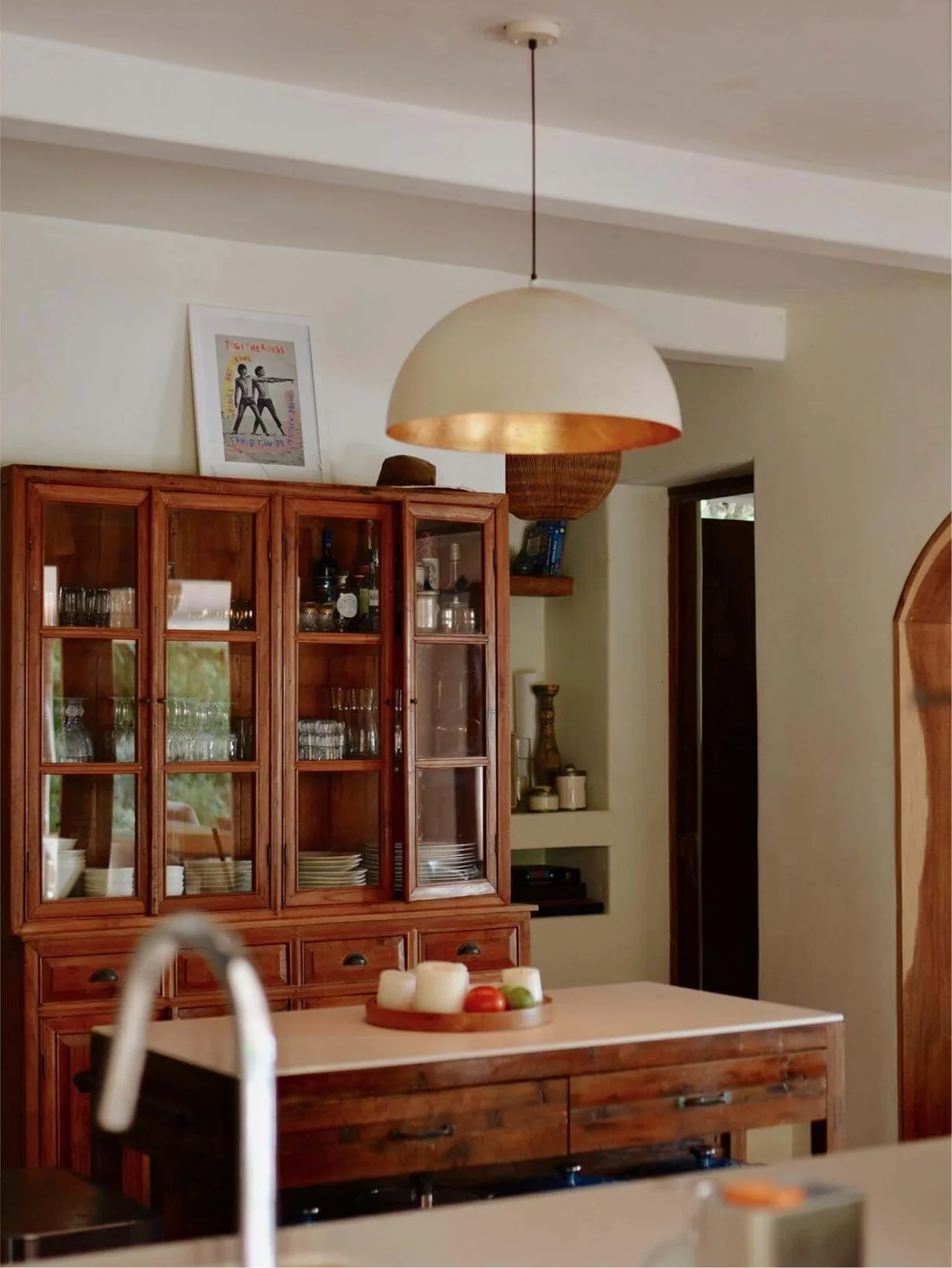 Kitchen with wooden cabinet filled with glassware and dishes, hanging ceiling light, a framed picture on the wall, and a white countertop with a tray of apples and candles.