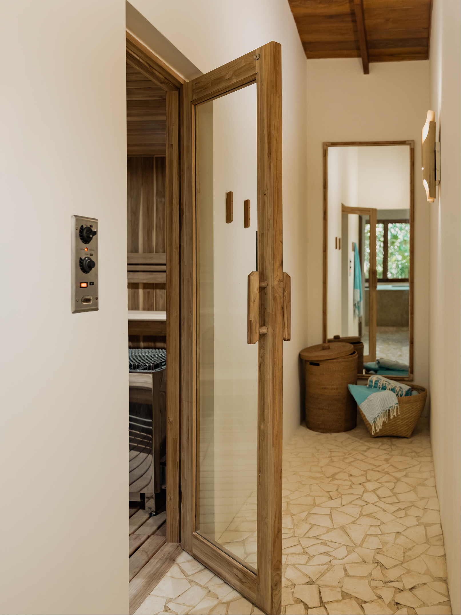 A hallway with white walls and a wooden ceiling. There is a full-length mirror with a wooden frame leaning against the wall. Next to it are two storage baskets, one round and the other rectangular, filled with towels. An open door reveals a room with