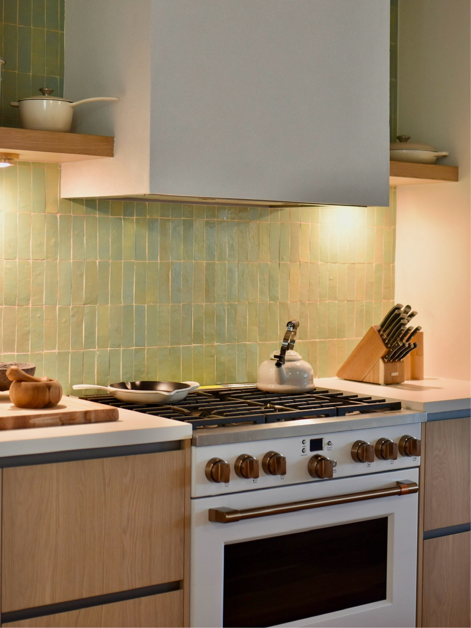 A modern kitchen stove with a white oven, wooden cabinet, and a green tiled backsplash. A kettle sits on the stove, and a knife block is on the counter.