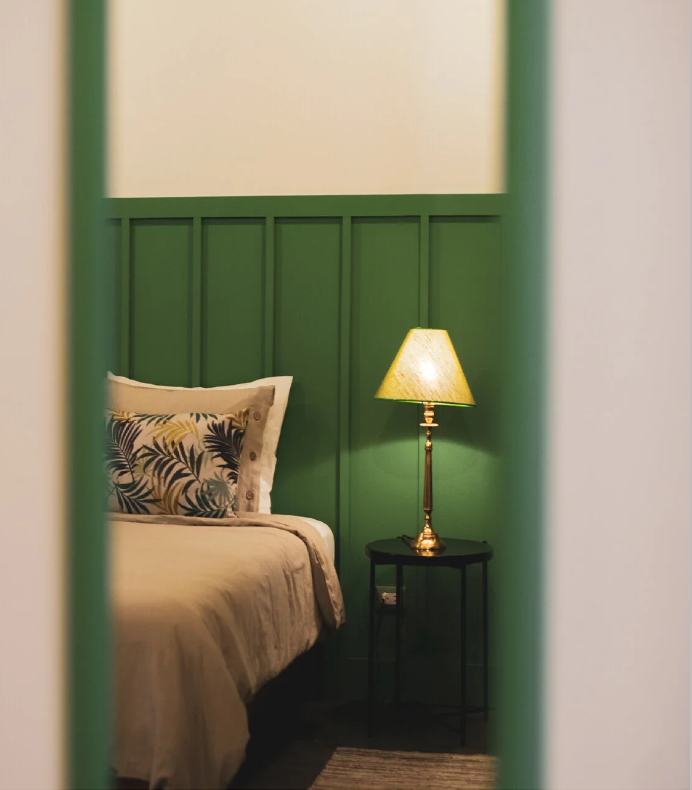 A bedroom viewed through an open doorway, featuring a bed with a patterned pillow, a black side table, a yellow-shaded lamp, and a green paneled headboard against a cream wall.