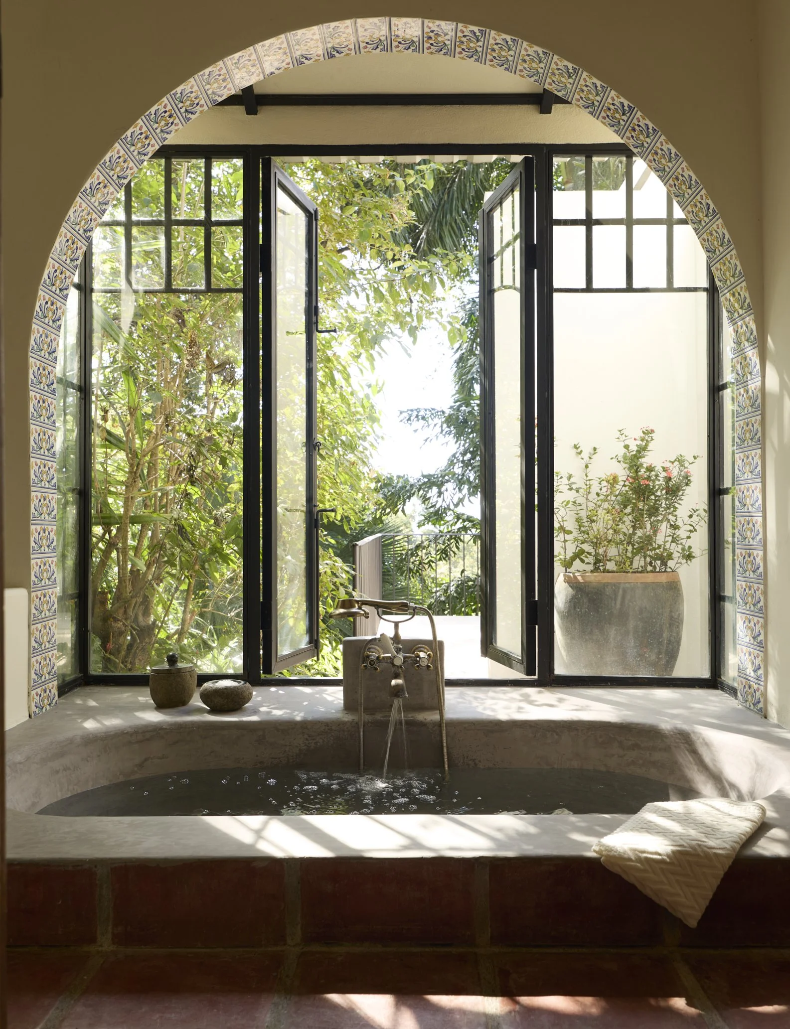 Inside view of a bathtub with water and a faucet, positioned beneath a large arched window with black frames, overlooking lush greenery outside. The window is decorated with patterned tiles along the arch, and there are two ceramic pots and a small stone on the tub's edge. A woven bath mat lies on the tile floor outside the tub.