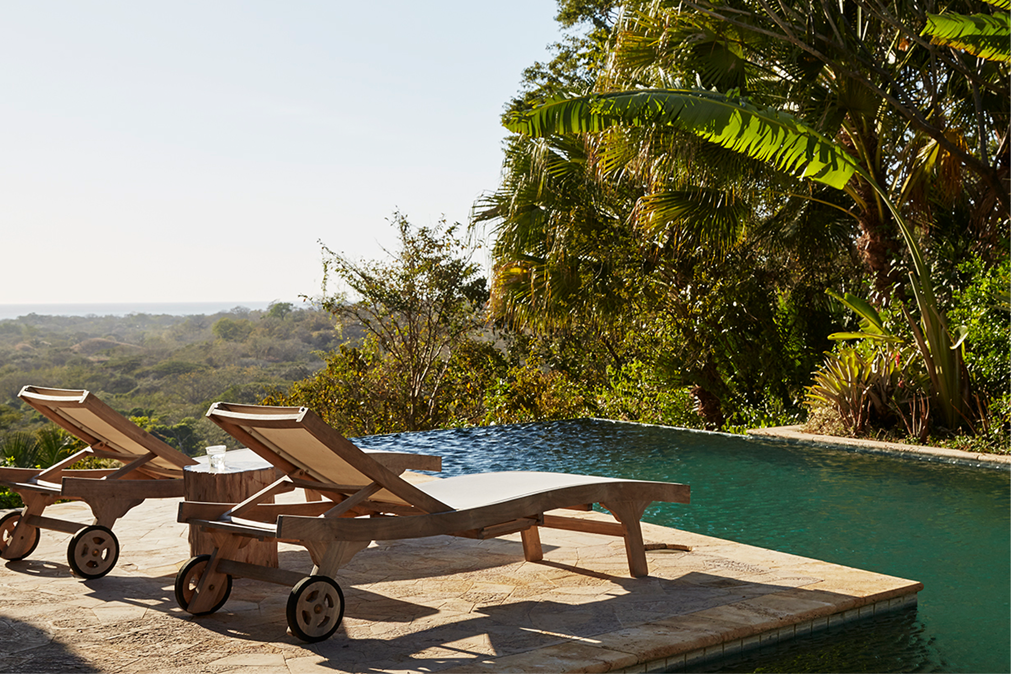 Poolside scene with lounge chairs, lush greenery, and trees in the background, under a clear sky.