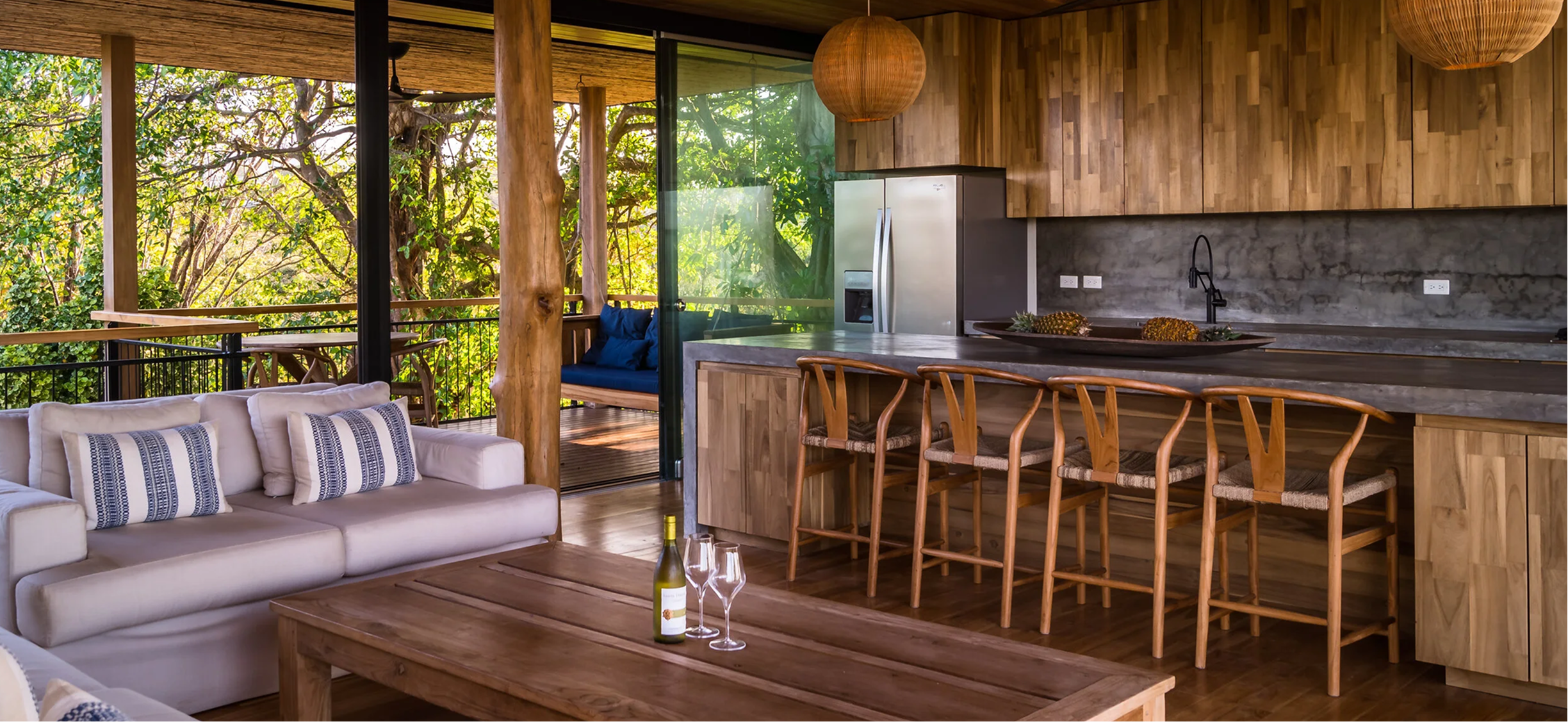 Open-concept living area with a cream sofa, wooden coffee table, and a view of a deck surrounded by trees. The kitchen features wooden cabinets, a concrete countertop, bar stools, and a stainless steel refrigerator. Large paper lanterns hang from the ceiling.