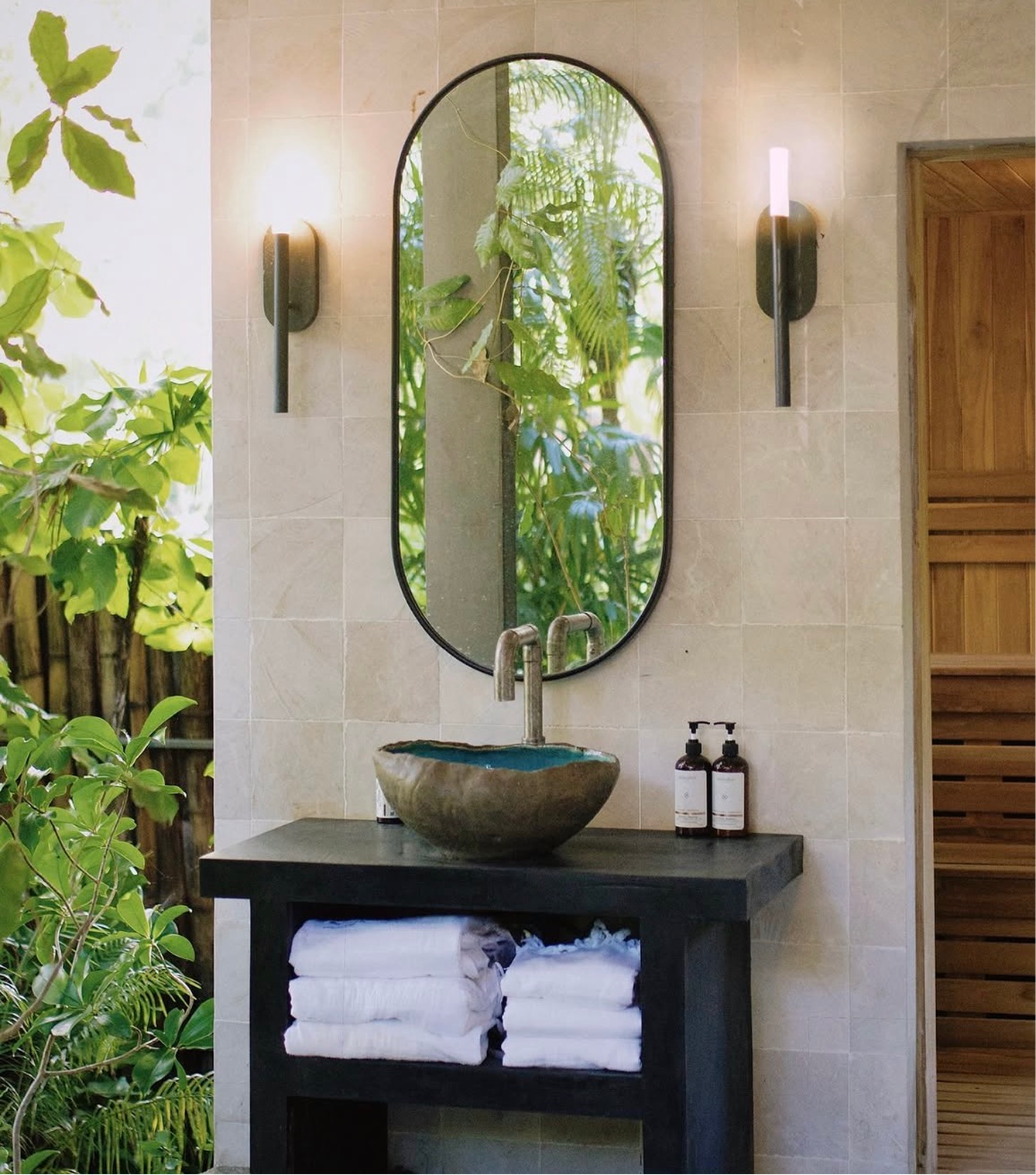 A bathroom vanity area with a stone sink, oval mirror, and black counter. Two black wall-mounted light fixtures flank the mirror, and under the counter are neatly folded white towels.