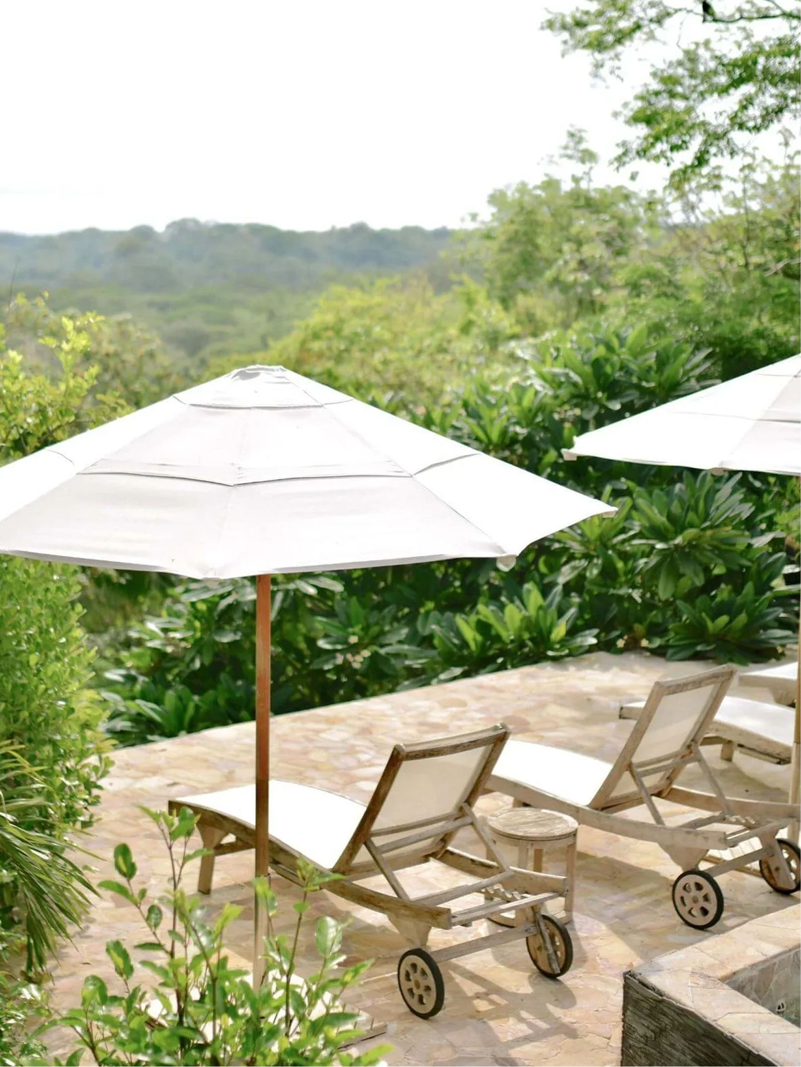 Two white patio umbrellas, two wooden lounge chairs, and a stone table on a patio with lush green bushes and trees in the background, overlooking a distant forested landscape.