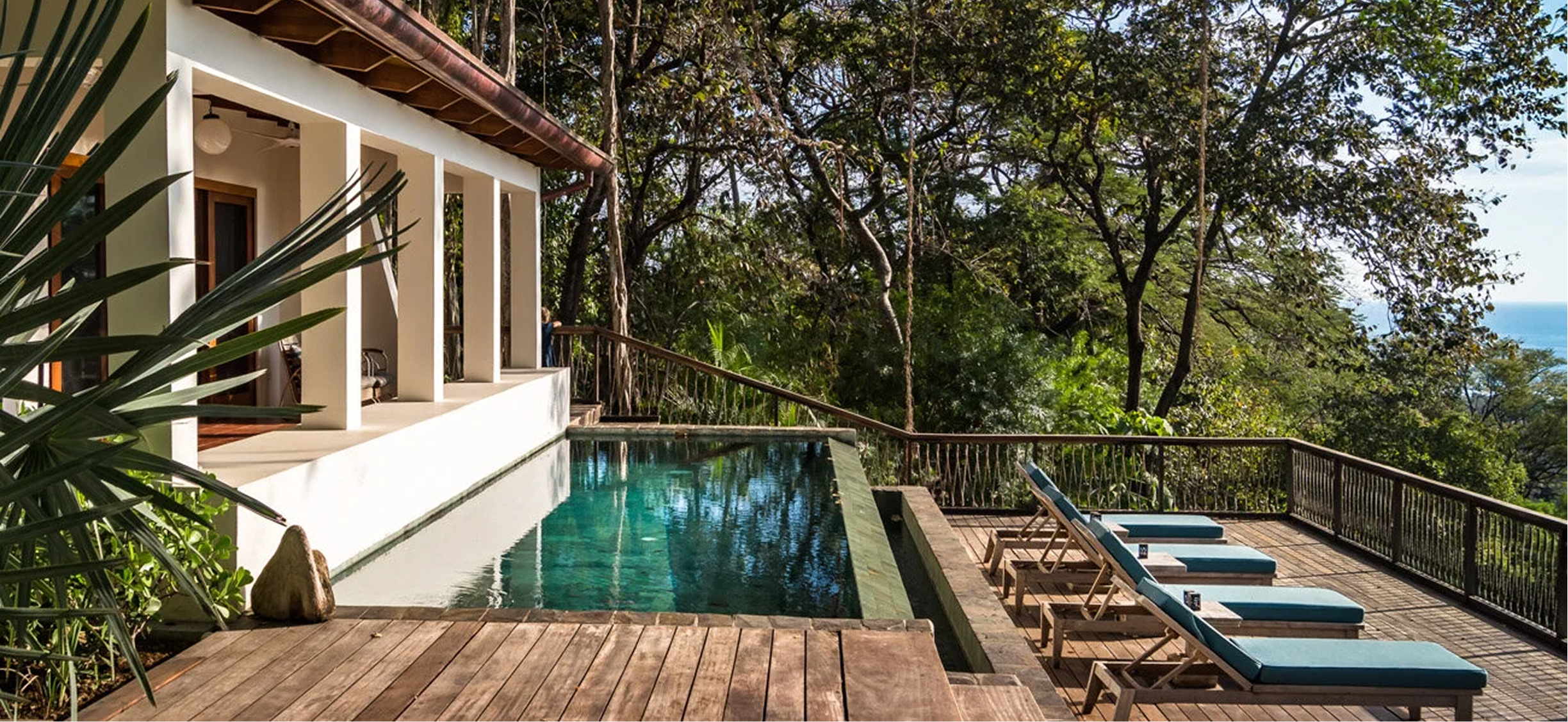 A wooden deck with lounge chairs beside a small infinity pool overlooking a lush forest with trees and a view of the ocean.