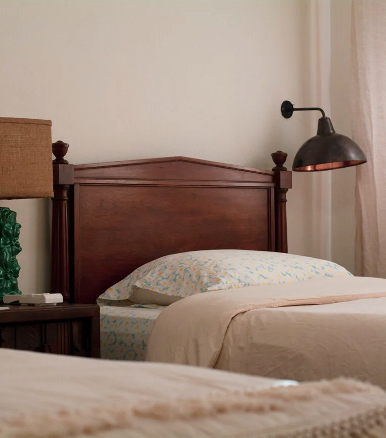 A cozy bedroom with a wooden headboard, a pillow with a floral pillowcase, a beige bedspread, a lamp attached to the wall above the headboard, a side table with a lamp and some items, a curtain on the right side, and soft lighting.