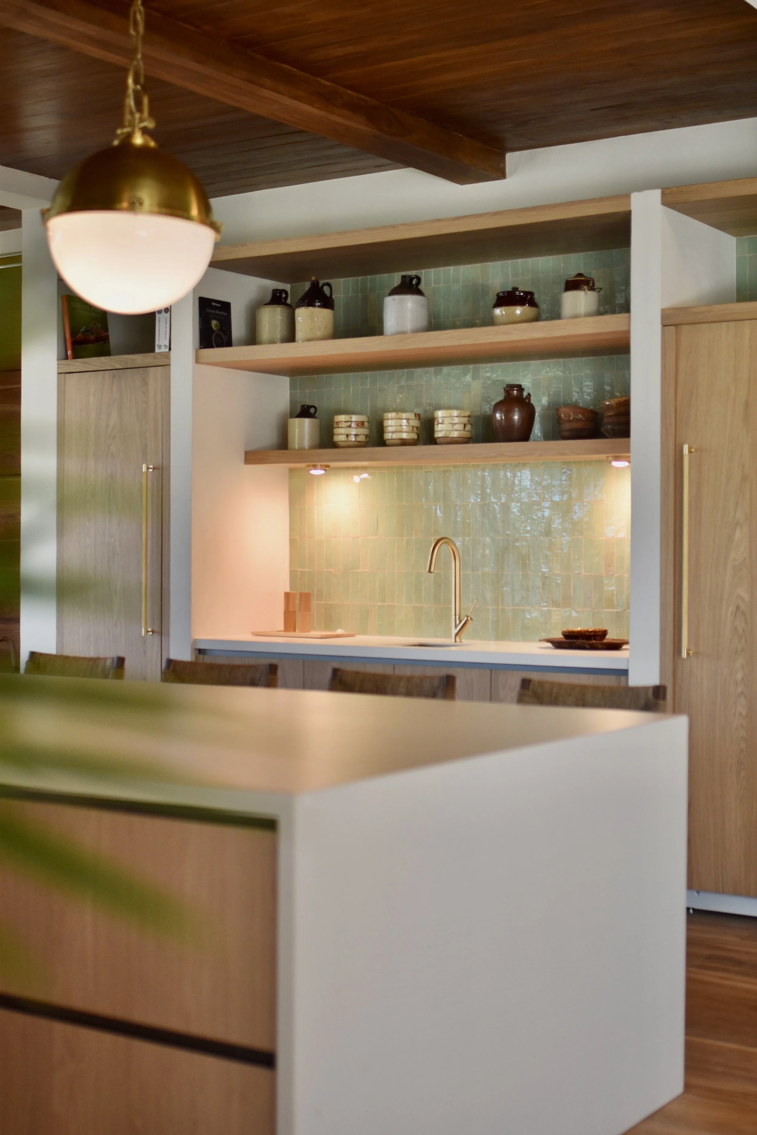 Modern kitchen with wooden ceiling and open shelves holding ceramic jars, bowls, and cookbooks. Green tiled backsplash and a white kitchen island with a gold faucet.