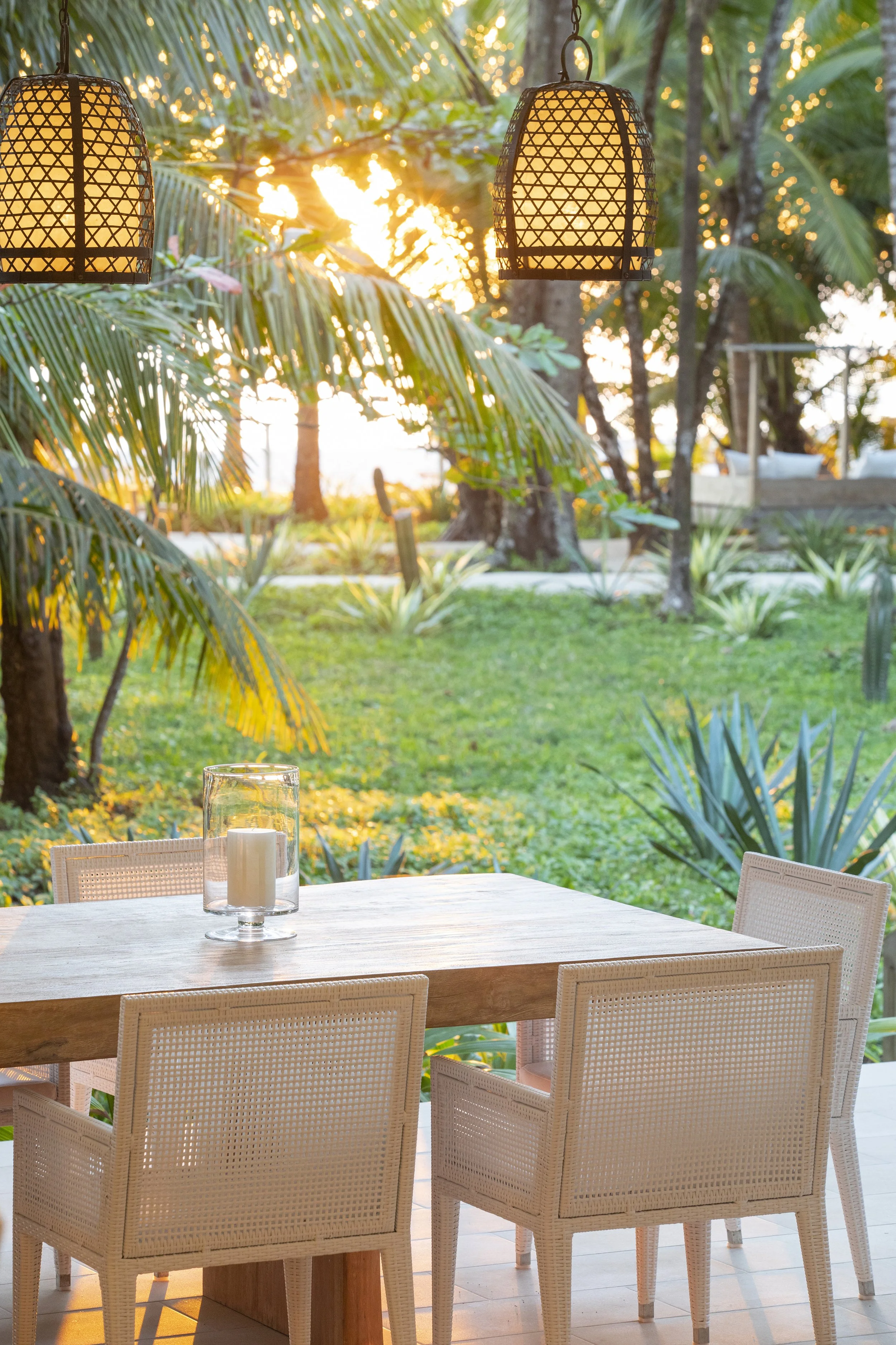 A dining table set outdoors with a candle in a glass holder on top, surrounded by wicker chairs, overlooking a lush garden with palm trees and greenery during sunset.