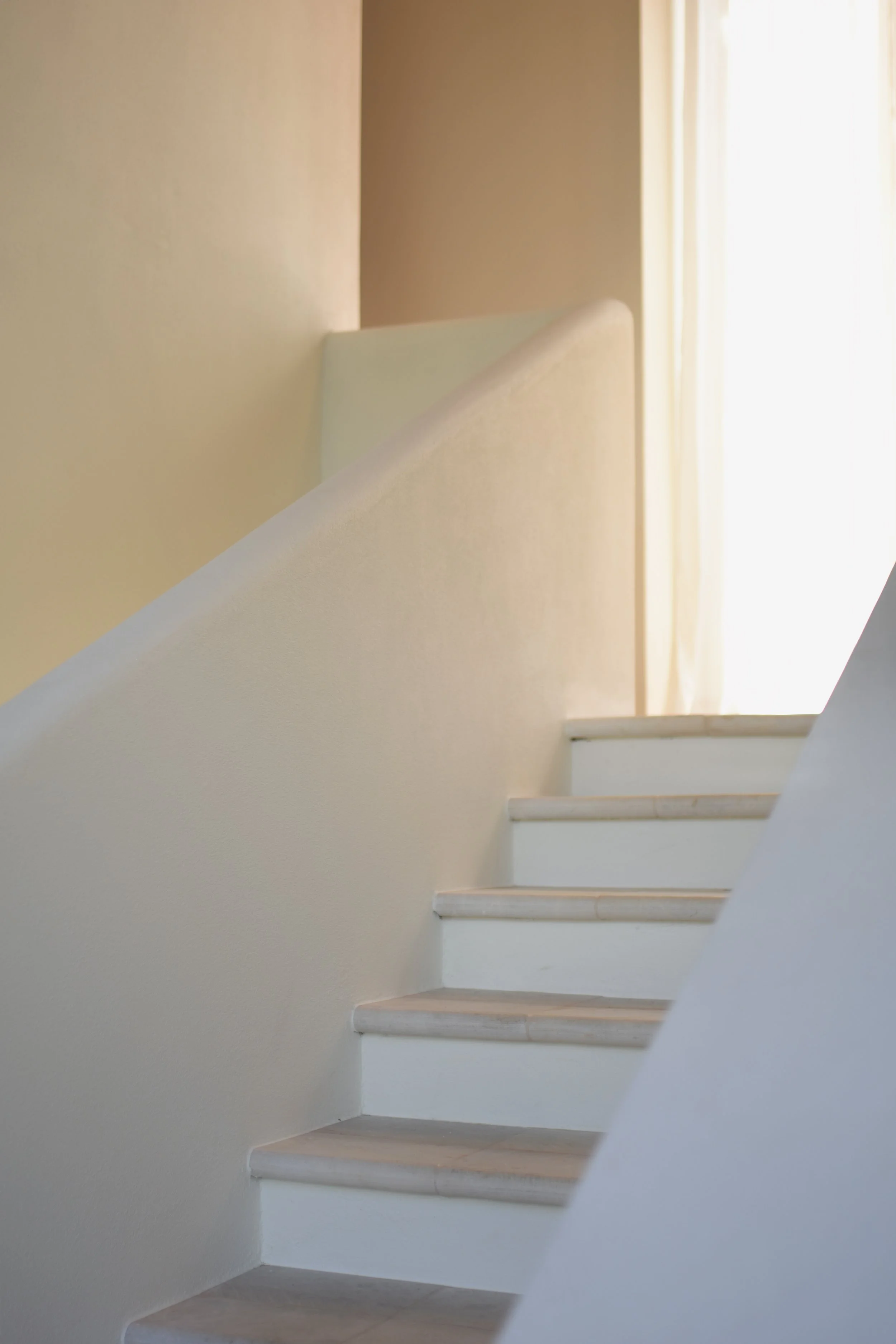 Interior view of a staircase with white steps and light-colored walls, illuminated by natural light from a window or doorway on the right side.