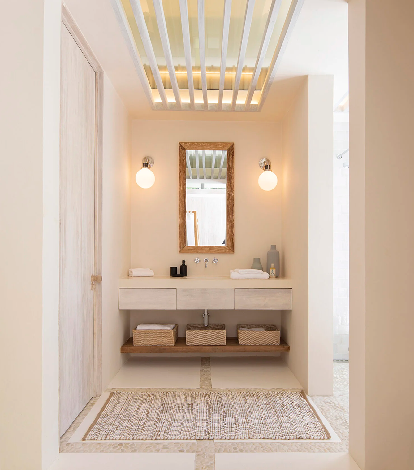 A minimalist bathroom with a floating vanity, a rectangular mirror with a wooden frame, and two frosted glass light fixtures on either side. Decor includes folded towels, ceramic vases, and a soap dispenser. The ceiling features a skylight with wooden beams, and the floor is covered with a woven rug.