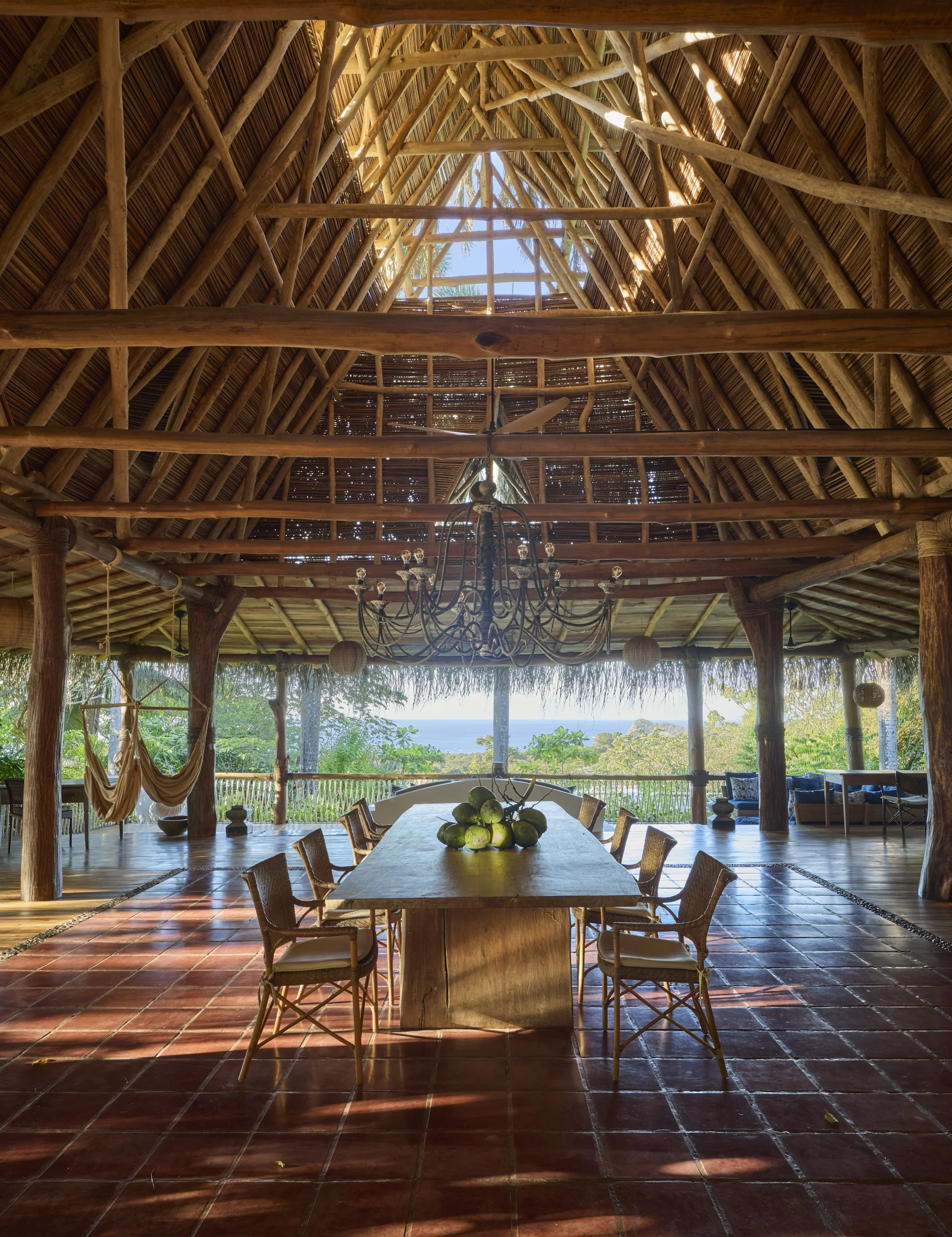 Open-air thatched roof structure with wooden beams, chandelier, dining table with green coconuts, and views of lush greenery and ocean in the background.
