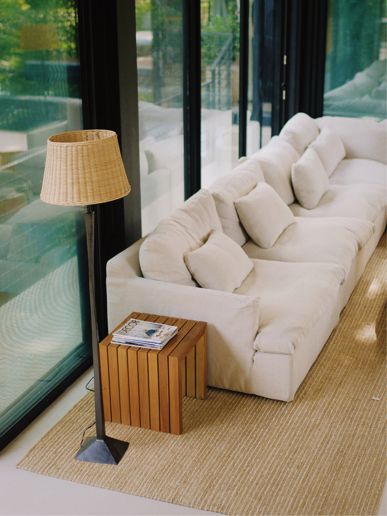 A living room with a cream-colored couch, beige cushions, a wooden side table with magazines, a floor lamp with a woven shade, large glass windows showing outdoor greenery, and a striped beige rug.
