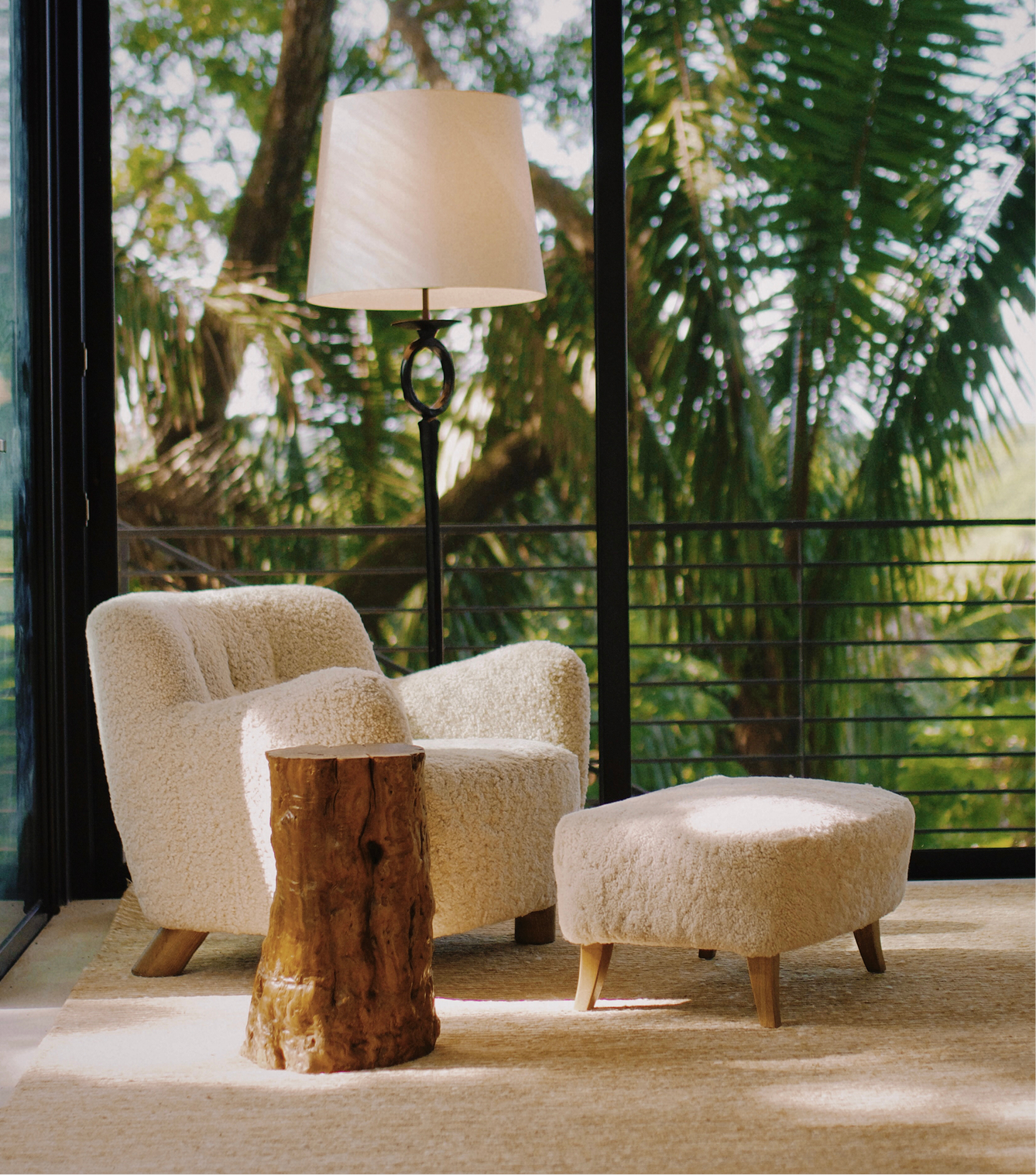 A cozy interior with a plush beige armchair, matching ottoman, and a small wooden stump side table, with a large window overlooking trees and greenery outside, and a tall floor lamp with a white lampshade.