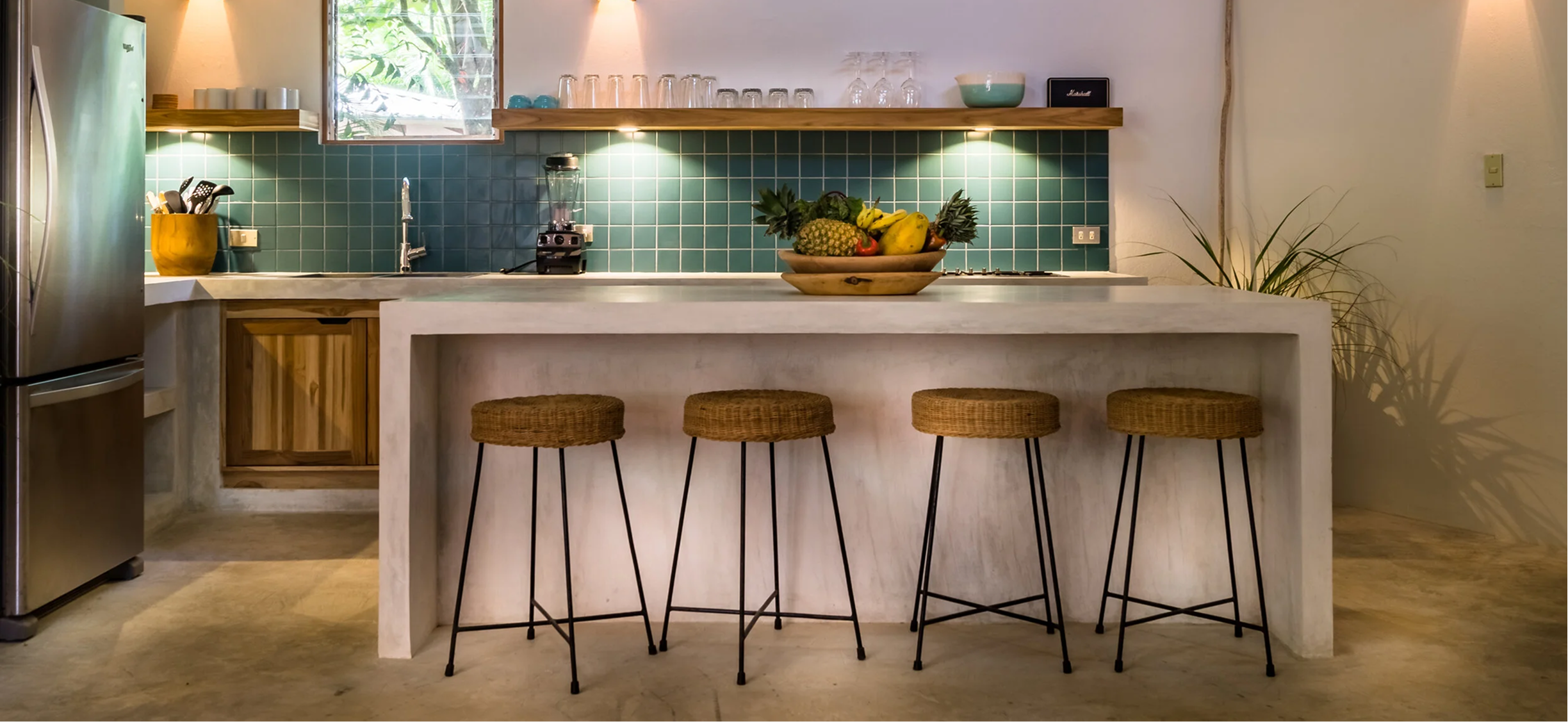 Modern kitchen with green tiled backsplash, white countertop, and wooden accents, featuring a bowl of fruit on the island and four woven bar stools.