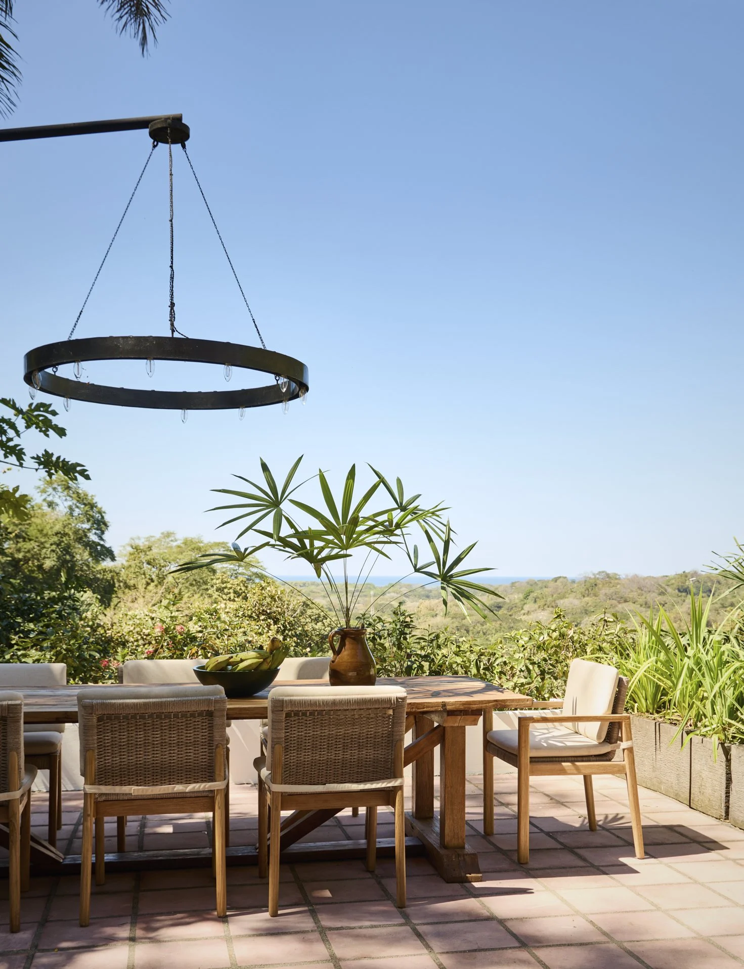 An outdoor patio dining area with a wooden table and chairs, a bowl of bananas, and a large potted plant, with a scenic view of trees and hills under a clear blue sky, and a circular chandelier hanging above.