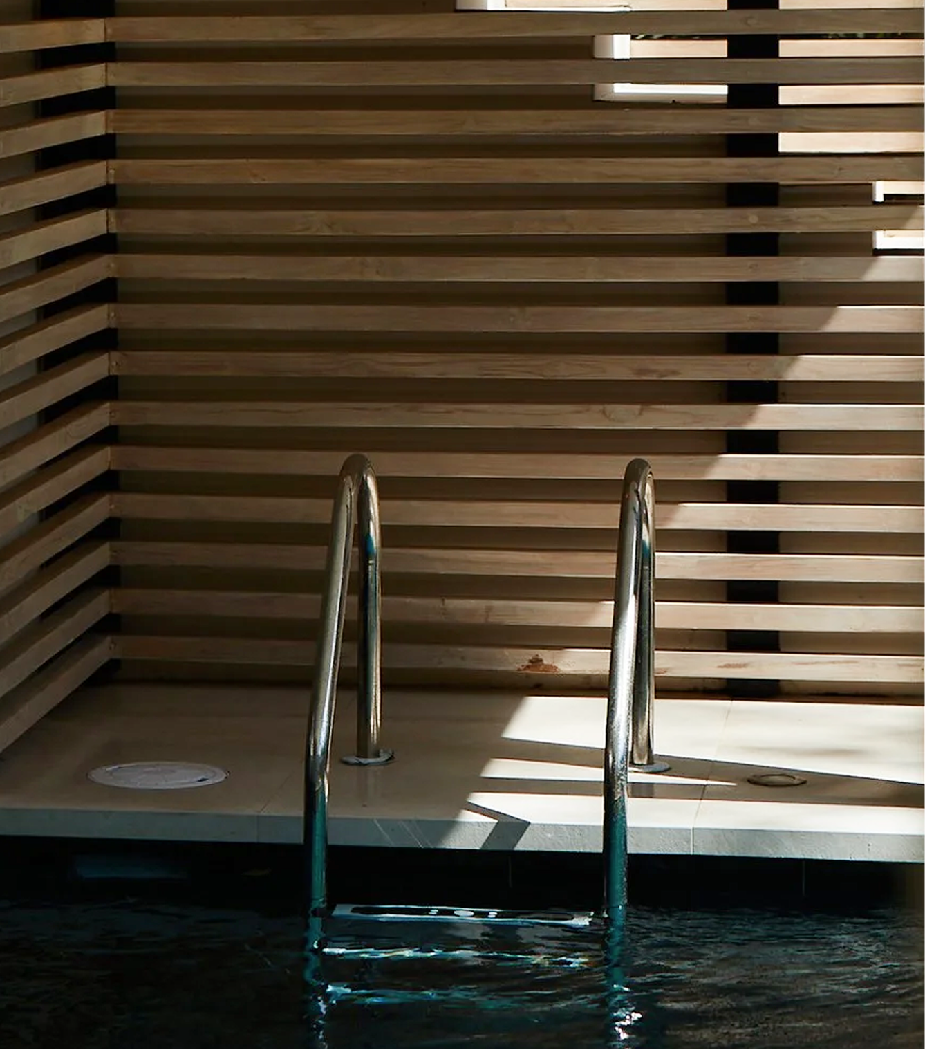 A swimming pool ladder in front of a wall made of horizontal wooden slats, with sunlight casting shadows on the wall.