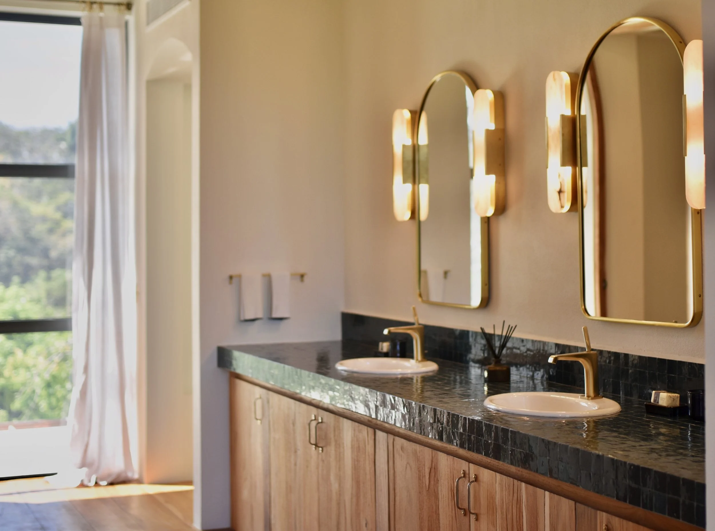 A bathroom with a black marble countertop, two sinks with gold fixtures, large mirrors, and vanity lights, wooden cabinets below, and a window with white curtains letting in natural light.