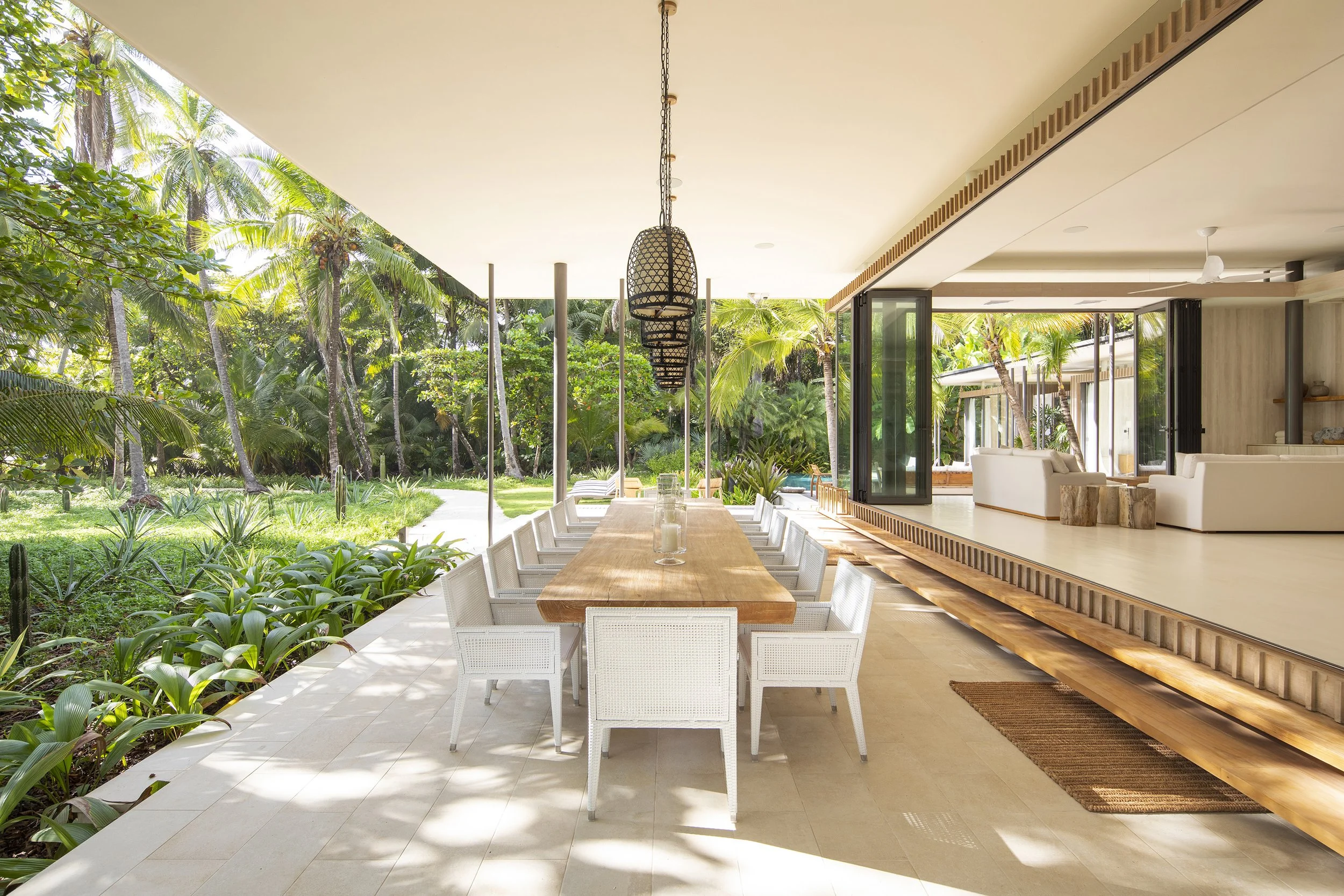 Outdoor dining area with a long wooden table surrounded by white chairs, overlooking a lush tropical garden with tall palm trees. The space is open to the garden with sliding glass doors and features modern decor, including hanging basket lights and a ceiling fan.