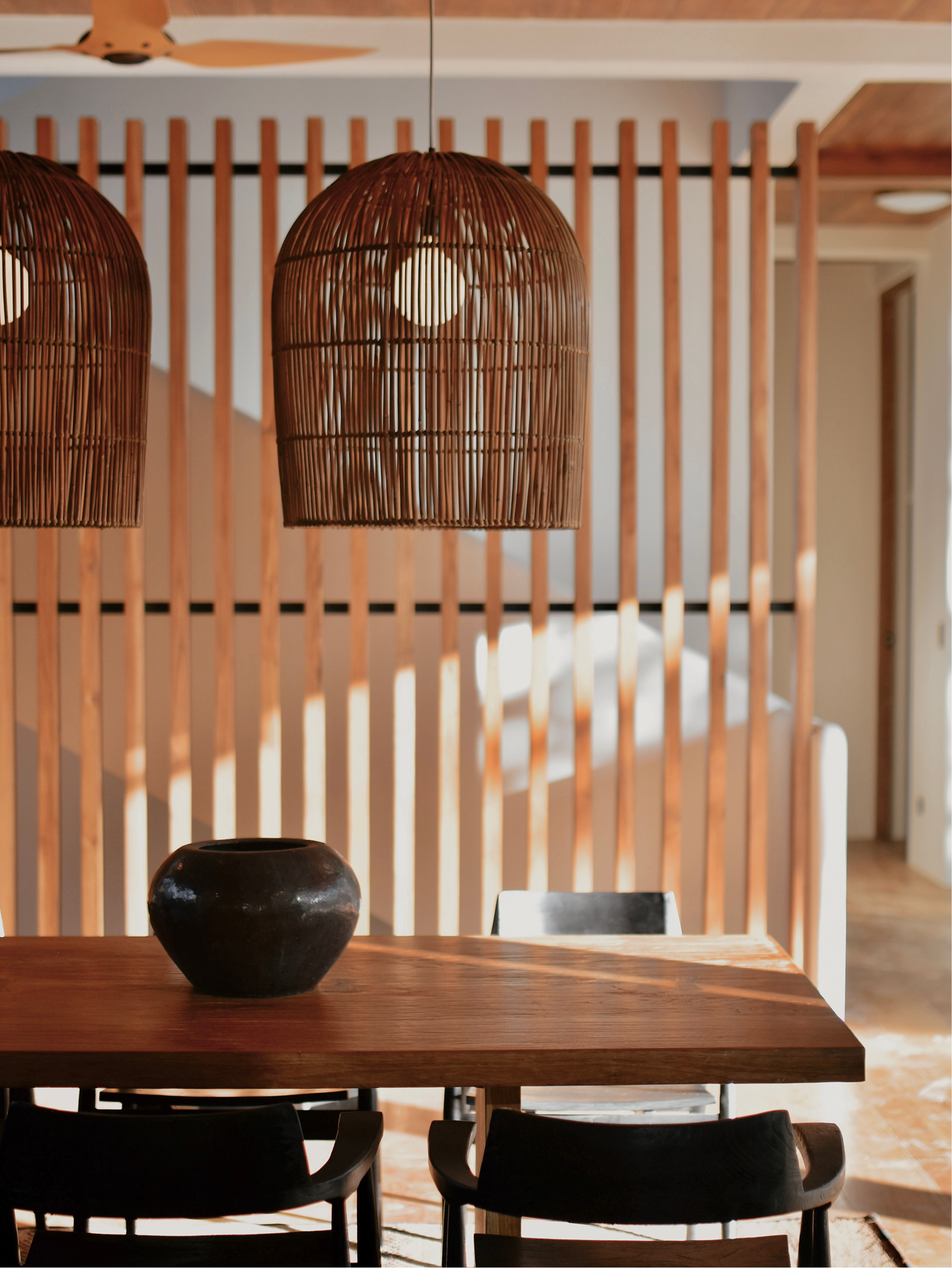 Interior of a room with a wooden table, black chairs, and two woven rattan pendant lights hanging from the ceiling. A black ceramic vase is placed on the table, and the background features vertical wooden slats and partial views of door frames and ce