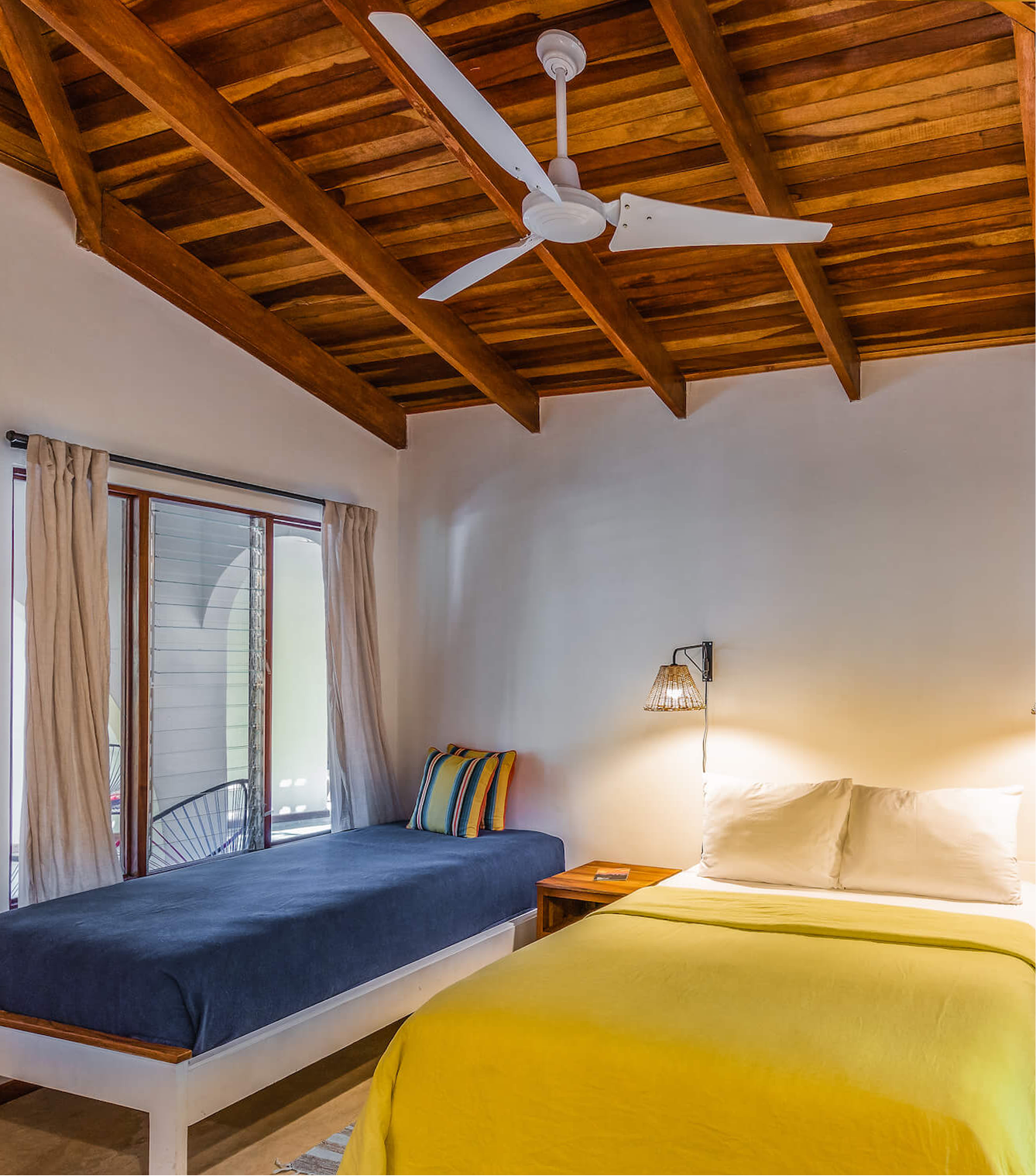 A bedroom with a wooden ceiling and a white ceiling fan. There are two beds: one with a dark blue bedspread and a striped pillow, and the other with a yellow bedspread and white pillows. A wooden nightstand with a book is between the beds. A wall-mounted lamp with a woven shade is beside the bed with the white pillows. There is a window with beige curtains, and sunlight is coming through.