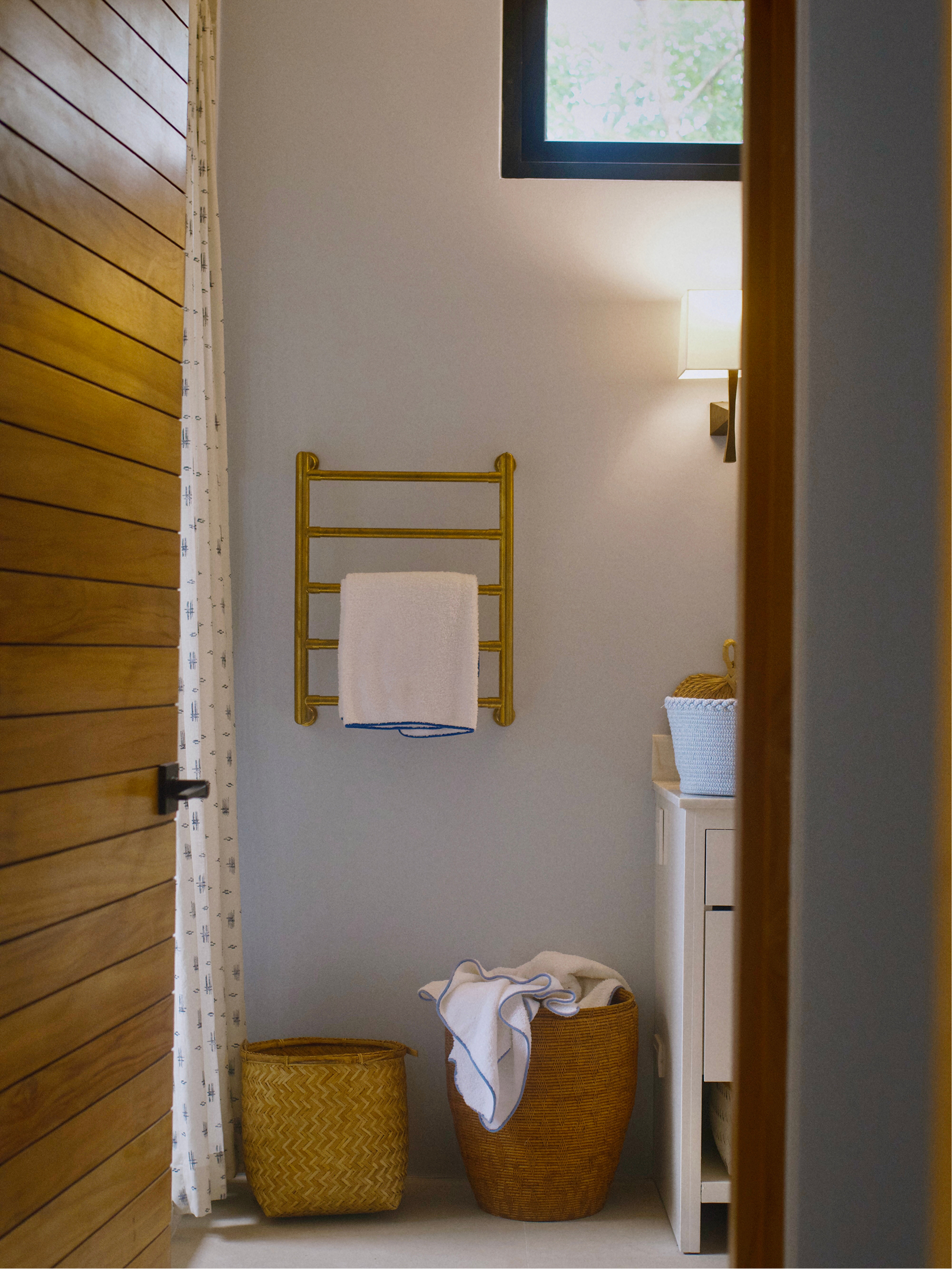 A bathroom corner with two woven baskets, a white towel hanging on a wall-mounted gold towel rack, a laundry basket with clothes, a bedside lamp on a small white cabinet, a window with green foliage views, and part of a wooden door and curtain.