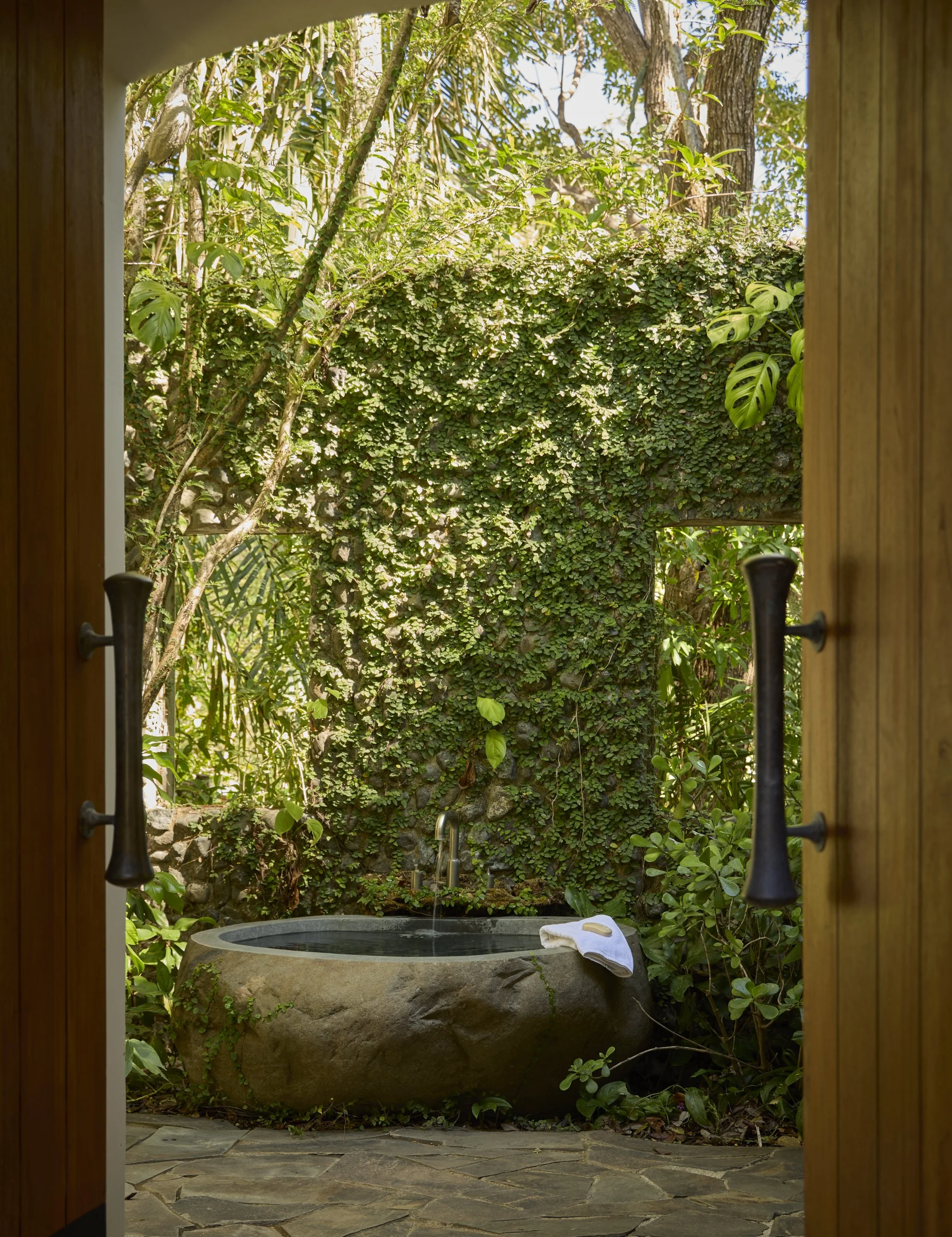Outdoor stone bathtub on a patio, surrounded by lush green foliage and trees, seen through a wooden doorway.