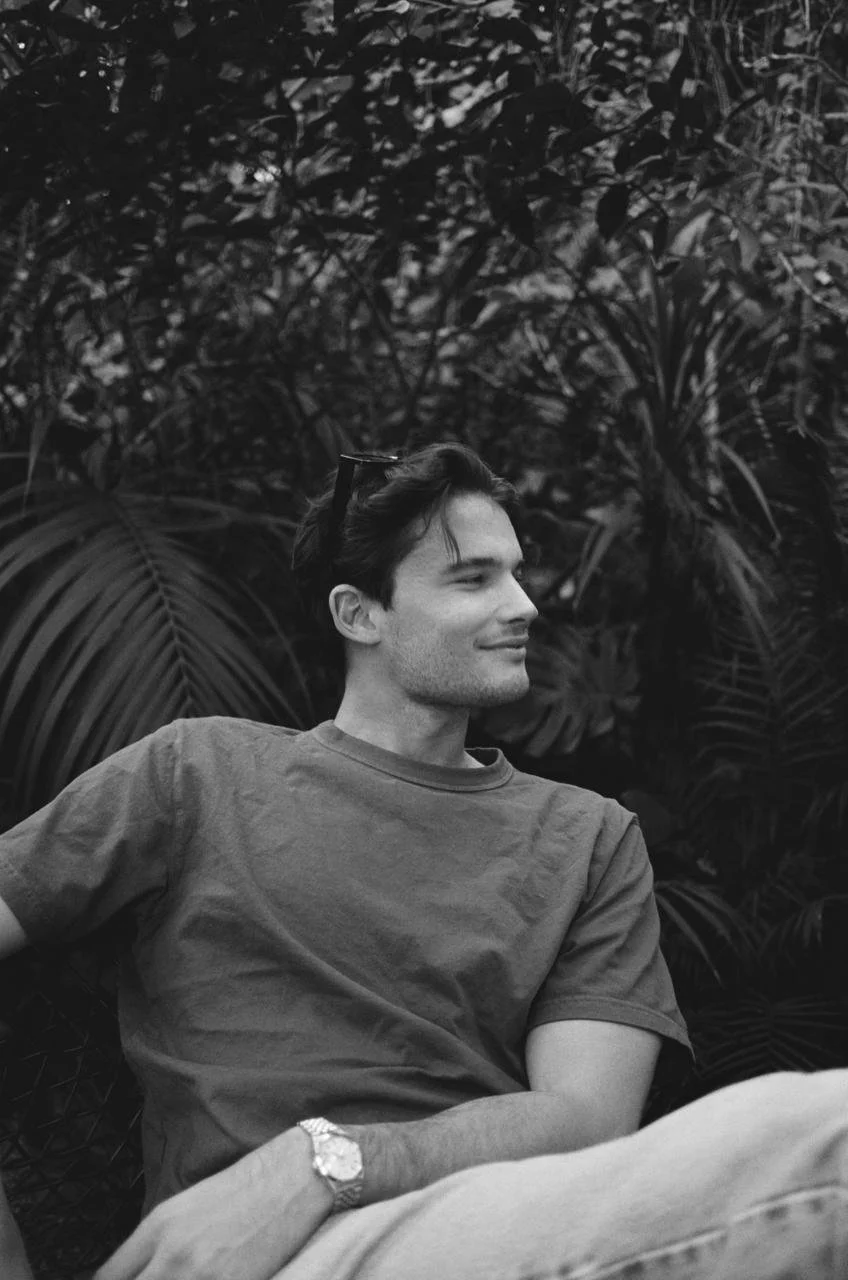 A young man sitting outdoors, smiling and looking to the side with sunglasses on his head, surrounded by lush tropical foliage.