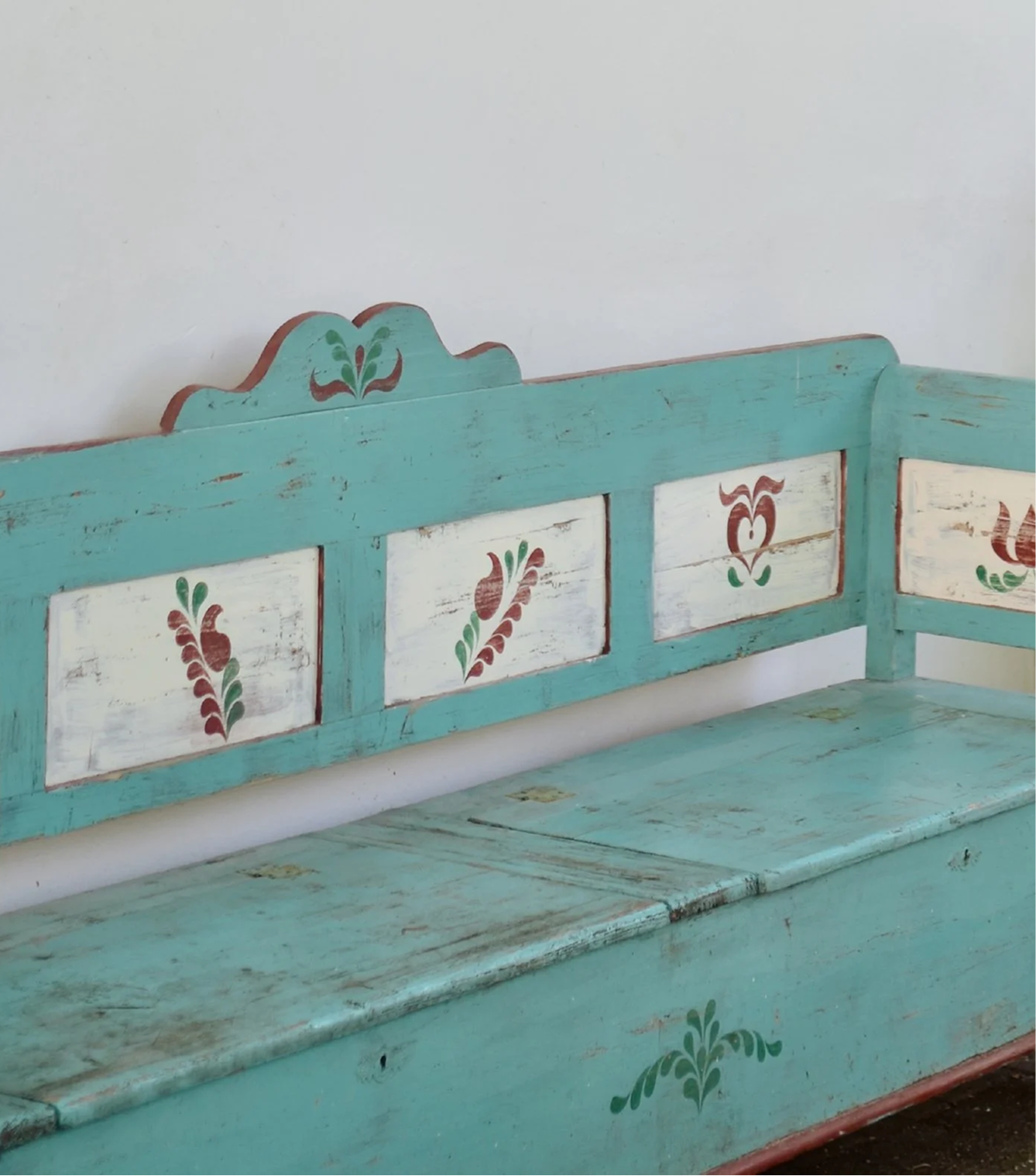 A vintage turquoise wooden bench with white panels decorated with floral patterns in red, green, and brown, placed against a plain white wall.