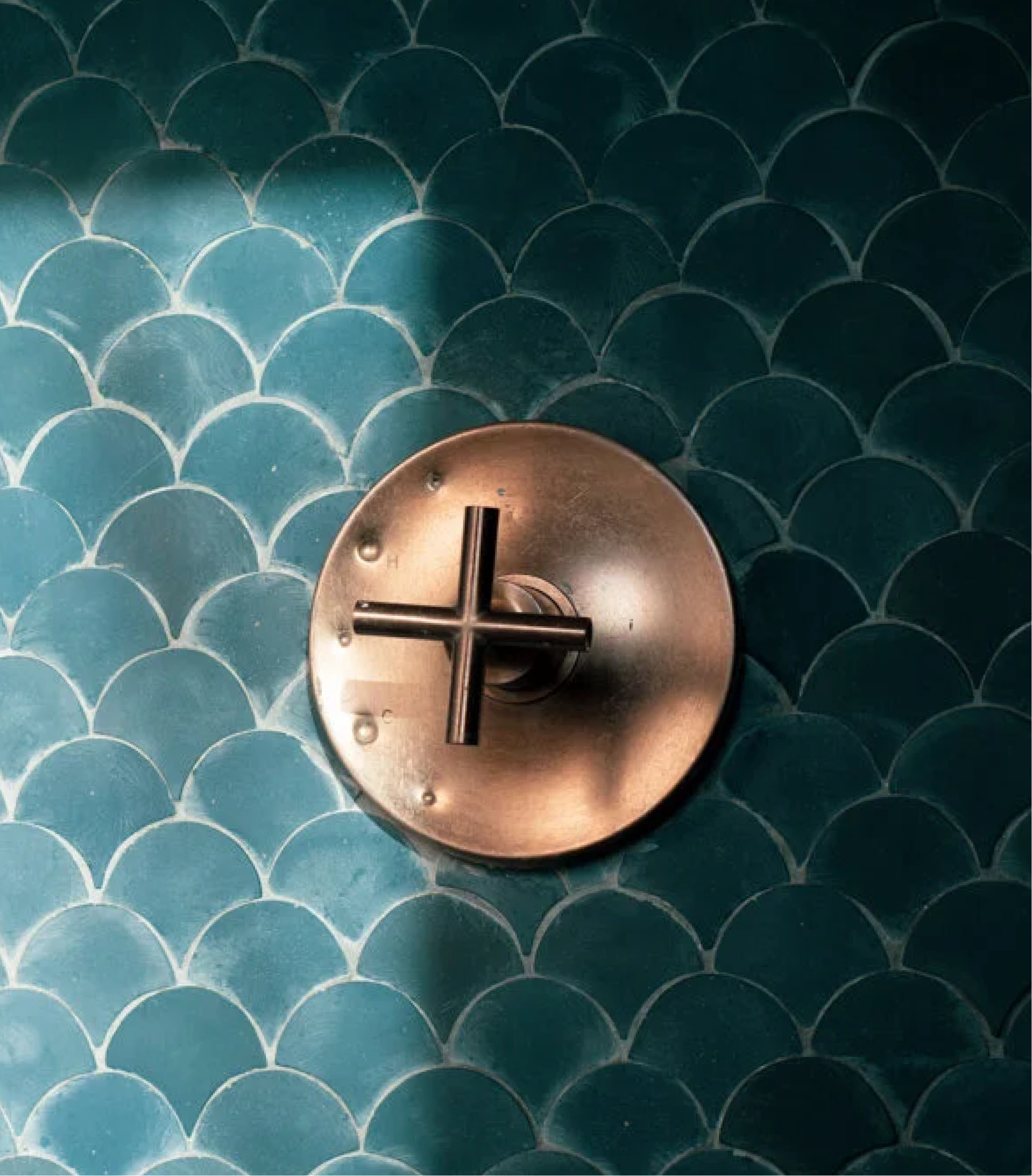 Close-up of a black metal object with a flat circular top and a metallic cross-shaped handle, sitting on a background of overlapping teal-blue scalloped tiles.