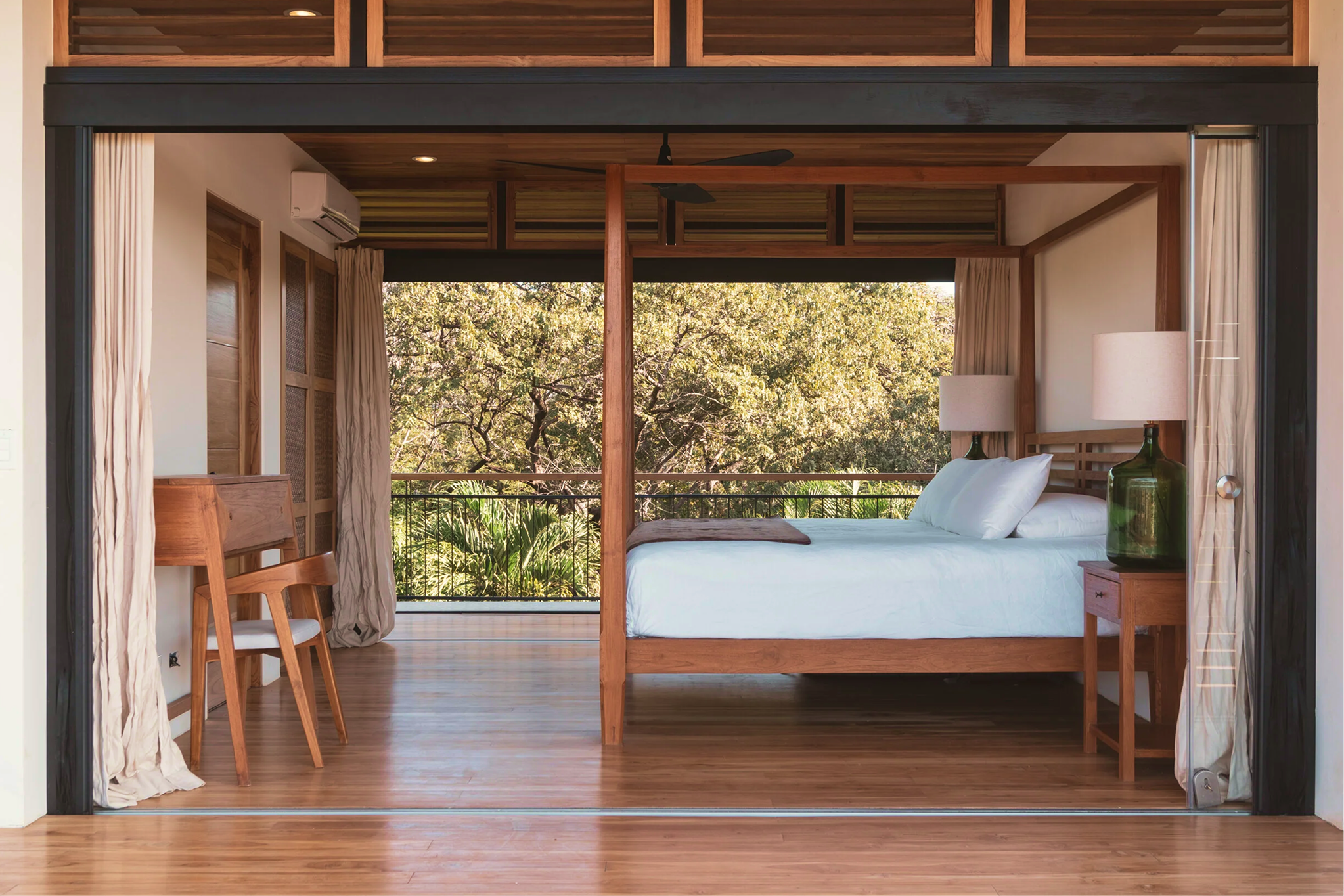 A bedroom with a wooden bed frame, white bedding, two nightstands with green lamps, large windows with curtains, and a view of trees outside.