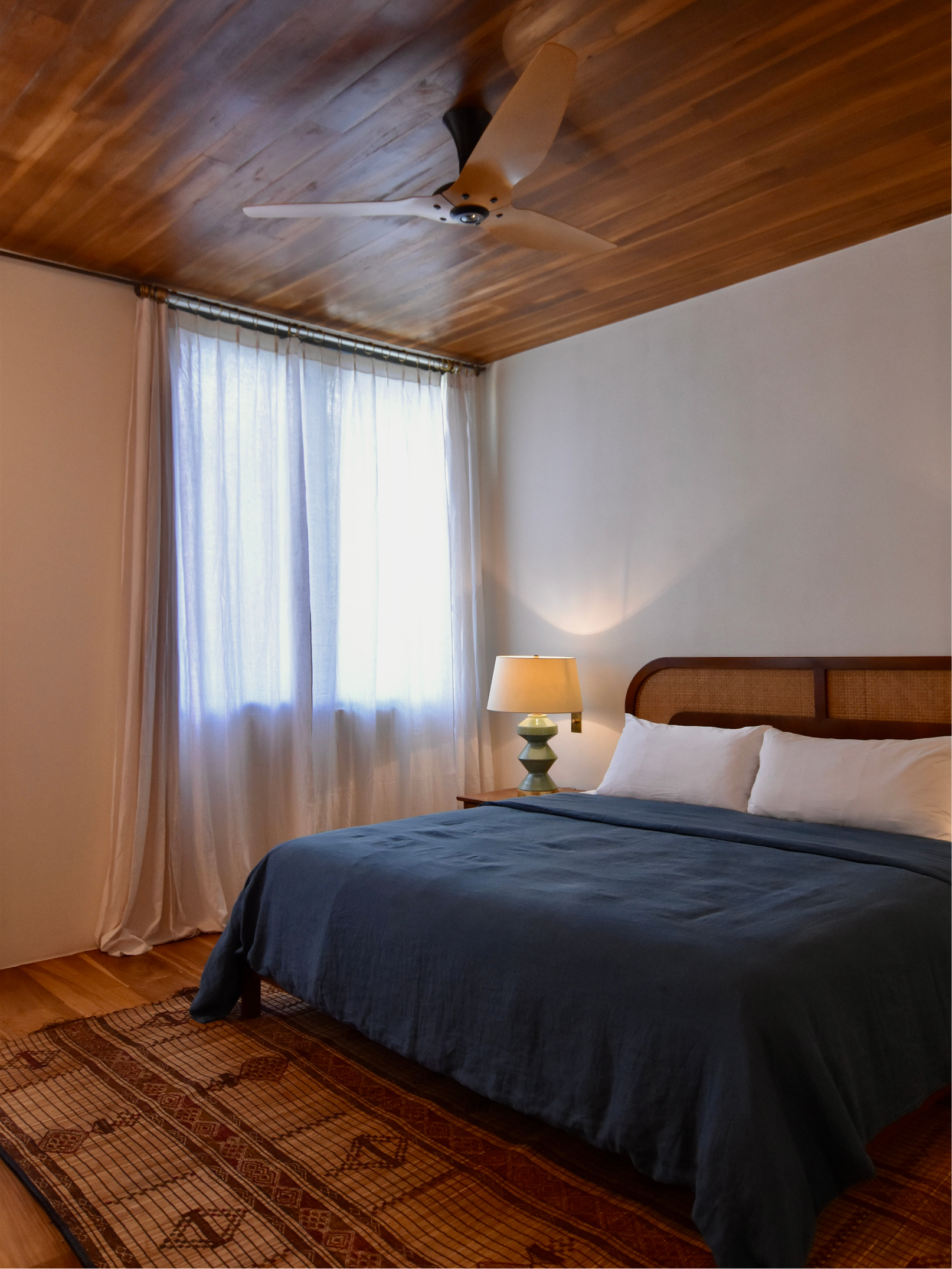 A bedroom with a bed covered in a dark blue bedspread, a bedside table with a lamp, a window with sheer white curtains, and a wooden ceiling with a ceiling fan.