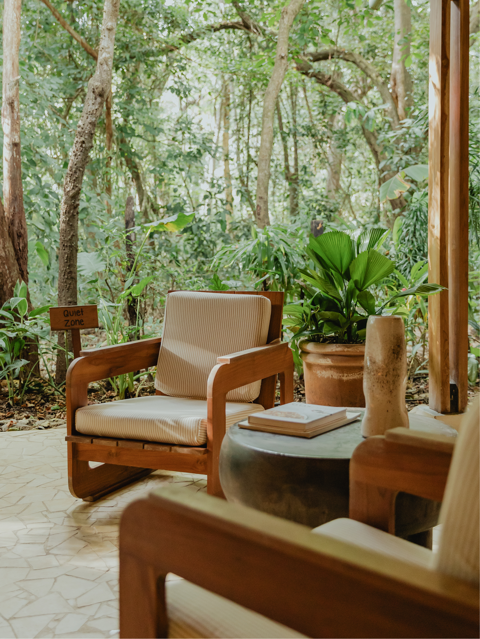 A cozy seating area with wooden chairs and a small round table in a lush, green forest setting. There are plants in pots, a book on the table, and a 'Quiet Zone' sign in the background.