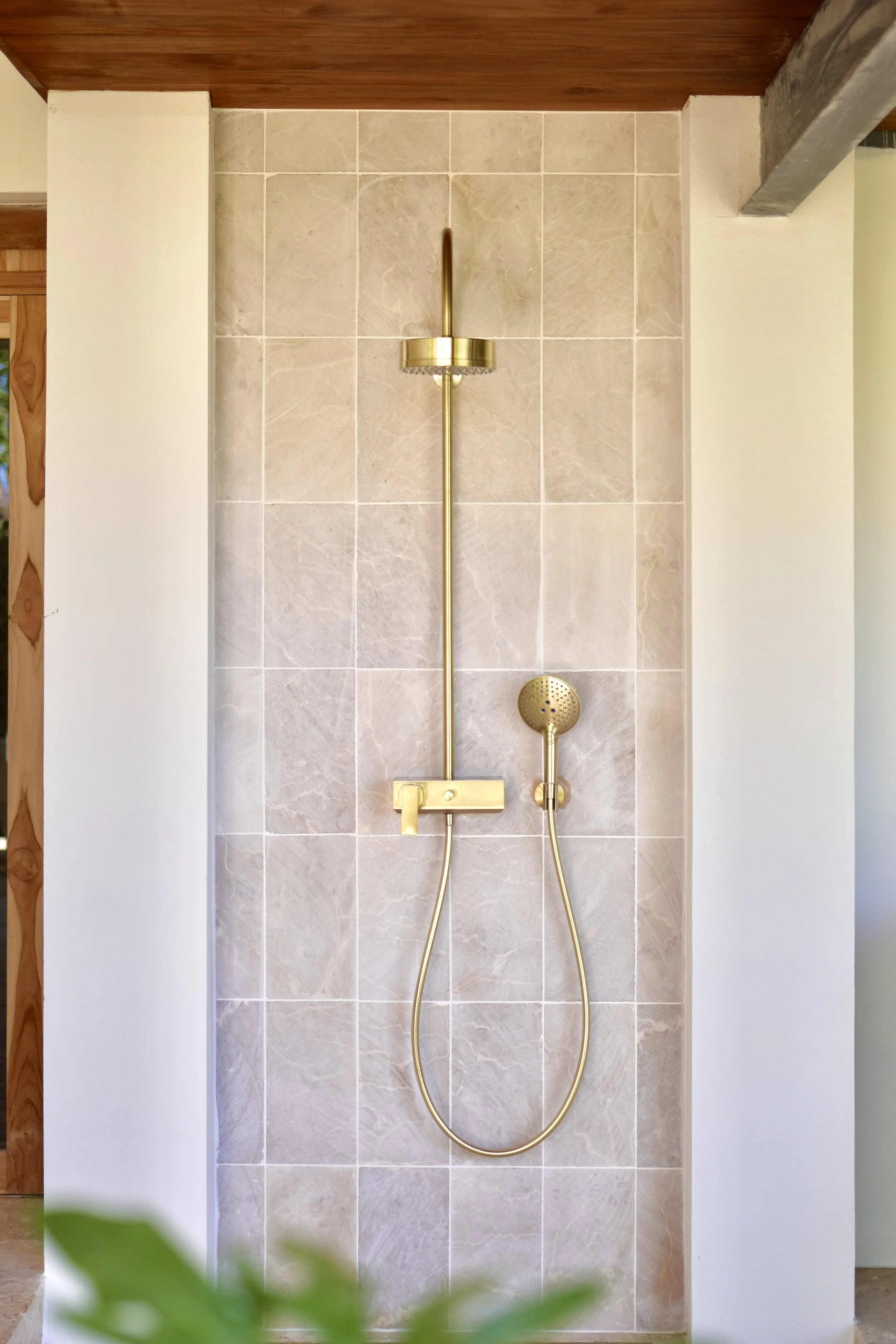 Modern shower with beige tile wall and gold fixtures, including a handheld showerhead and overhead rain shower, in a minimalist design.