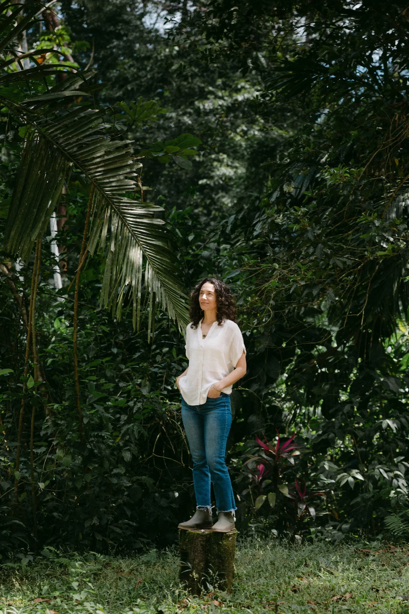 Alexis Lowenstein standing on a tree stump in a lush, green forest. She is wearing a white blouse, blue jeans, gray boots, and has her hands in her pockets, gazing off to the side.