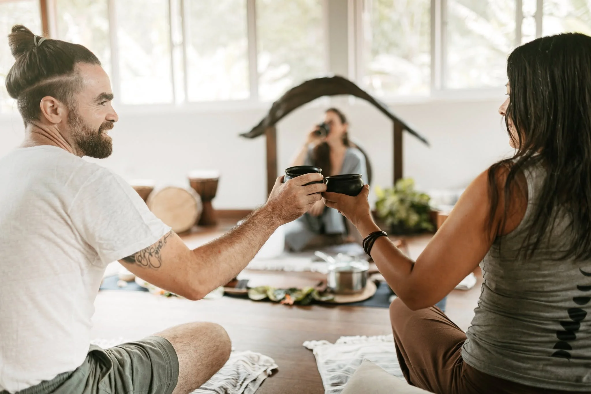 A man and woman with dark hair and tattoo on arm clink cups of cacao while sitting on the floor in a bright, airy room with large windows. They are participating in a cacao ceremony at the Sattva Land Retreat Center in Belize.