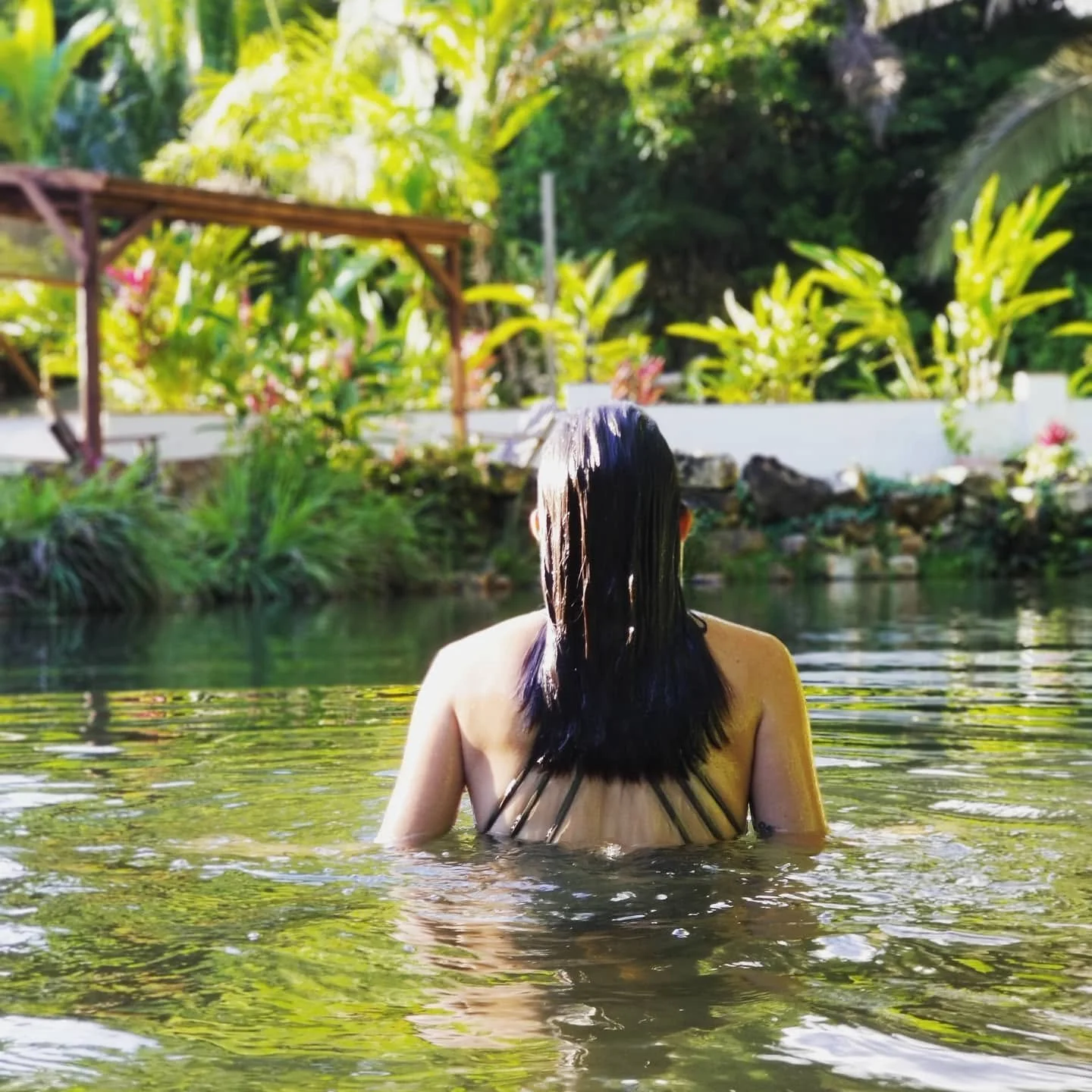 Woman with wet dark hair taking a bath in a natural pool surrounded by lush green plants and trees at the Sattva Land Retreat Center in Belize.