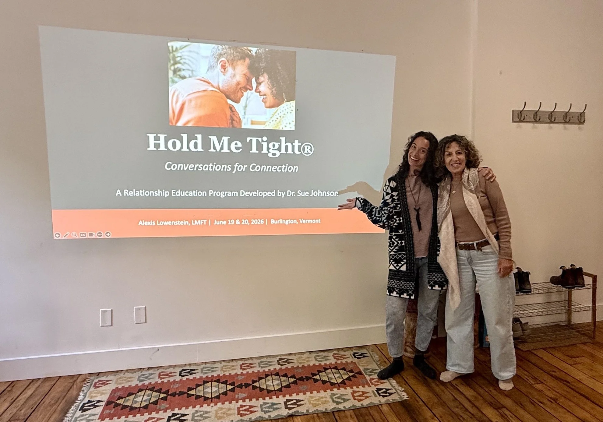 Alexis Lowenstein and another woman at a Hold Me Tight Workshop for Couples, standing indoors next to a wall with a projection screen.